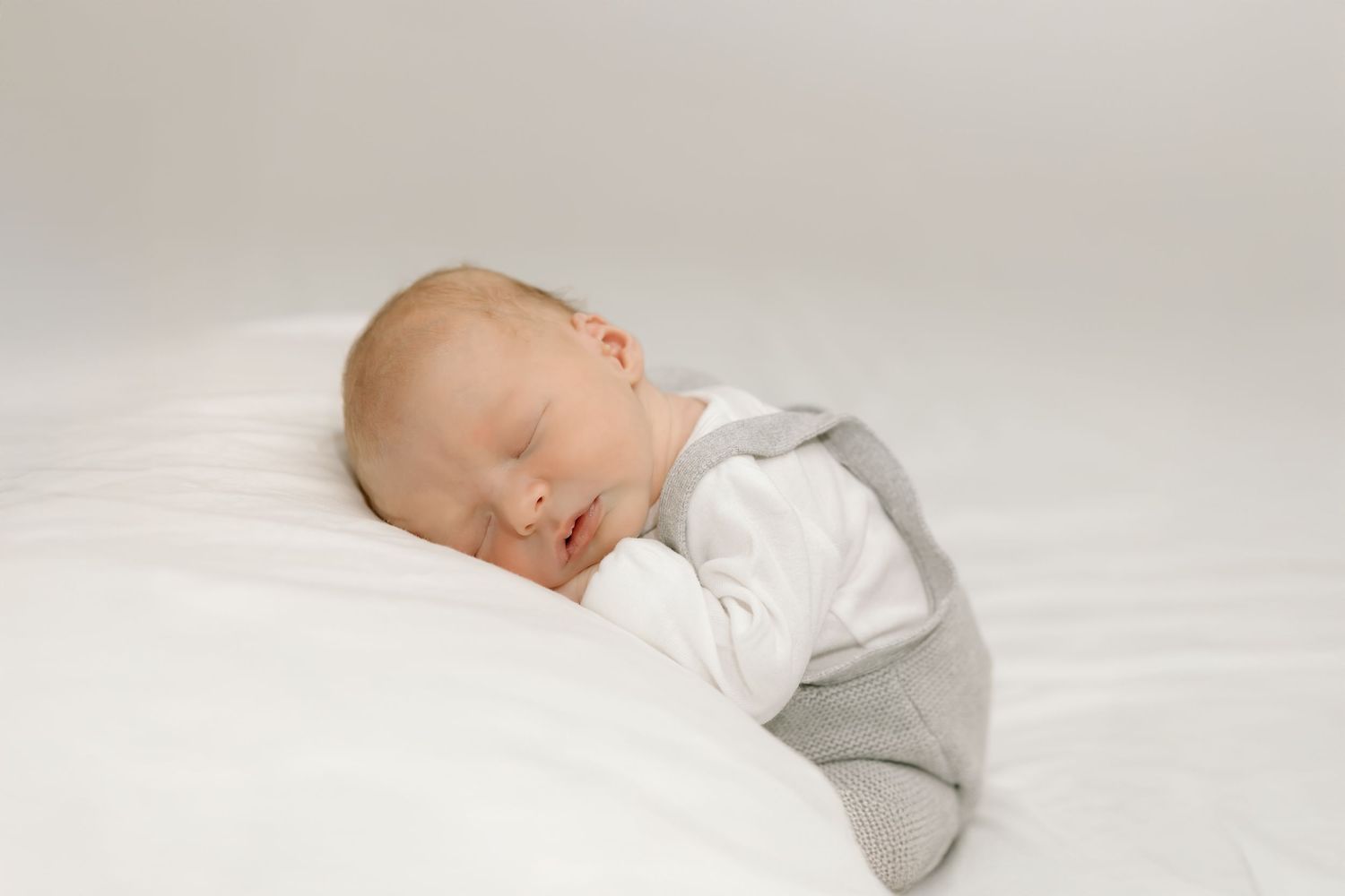 A sleeping baby wearing gray overalls and white shirt rests peacefully on white bedding.