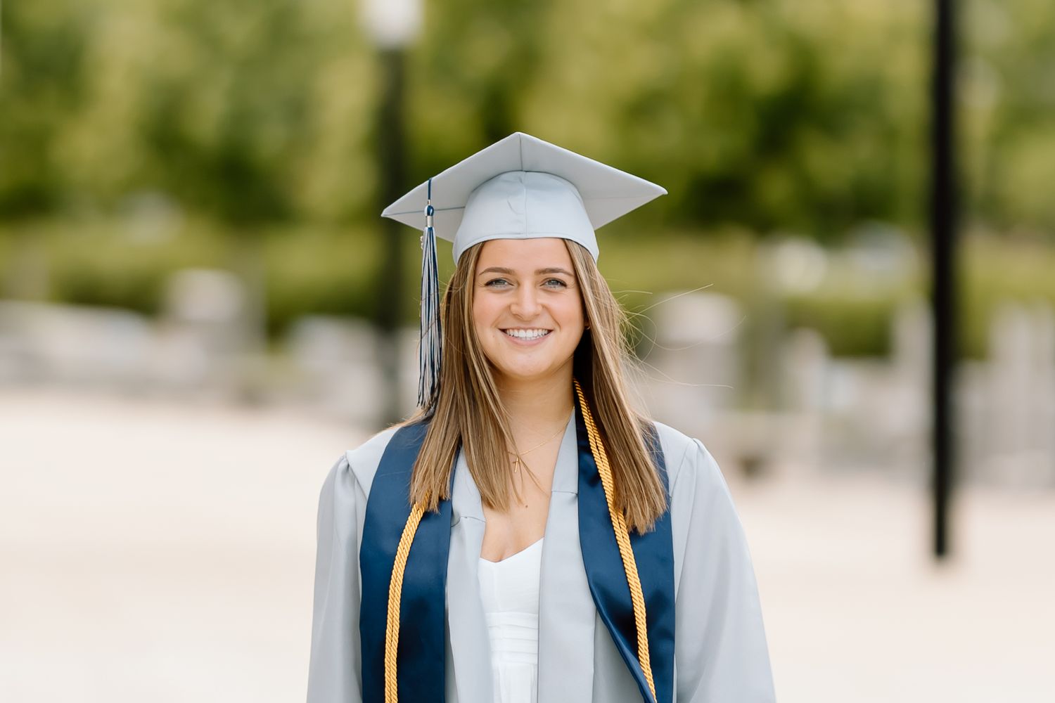 Graduate wearing a white cap and gown stands outdoors with a bright smile on a sunny day.