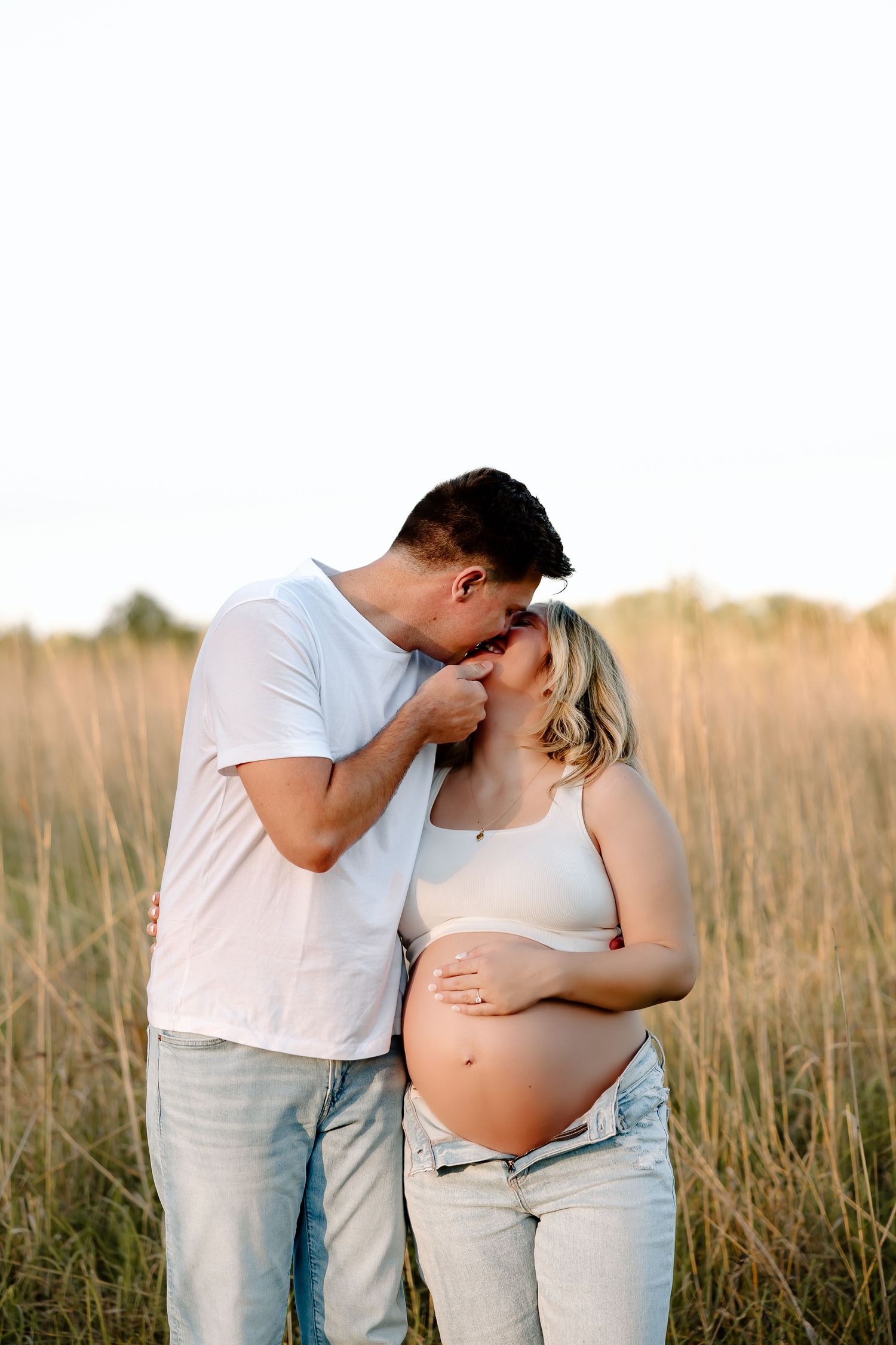 Couple shares intimate moment in golden field during sunset maternity photoshoot.