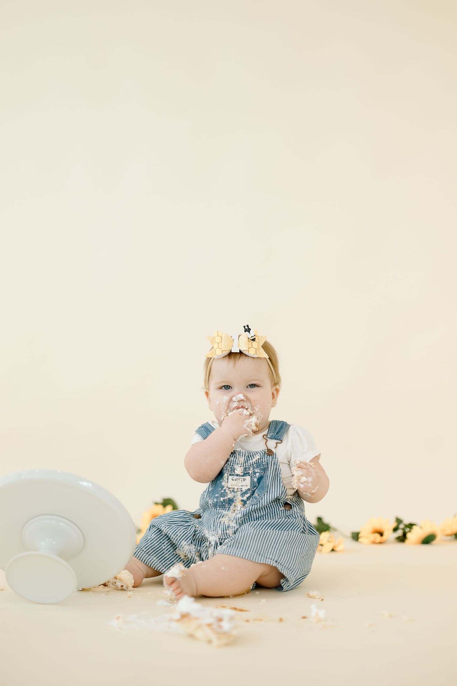 Baby in denim overalls sits on cream backdrop with birthday cake smash photo session showing messy frosting and fun.
