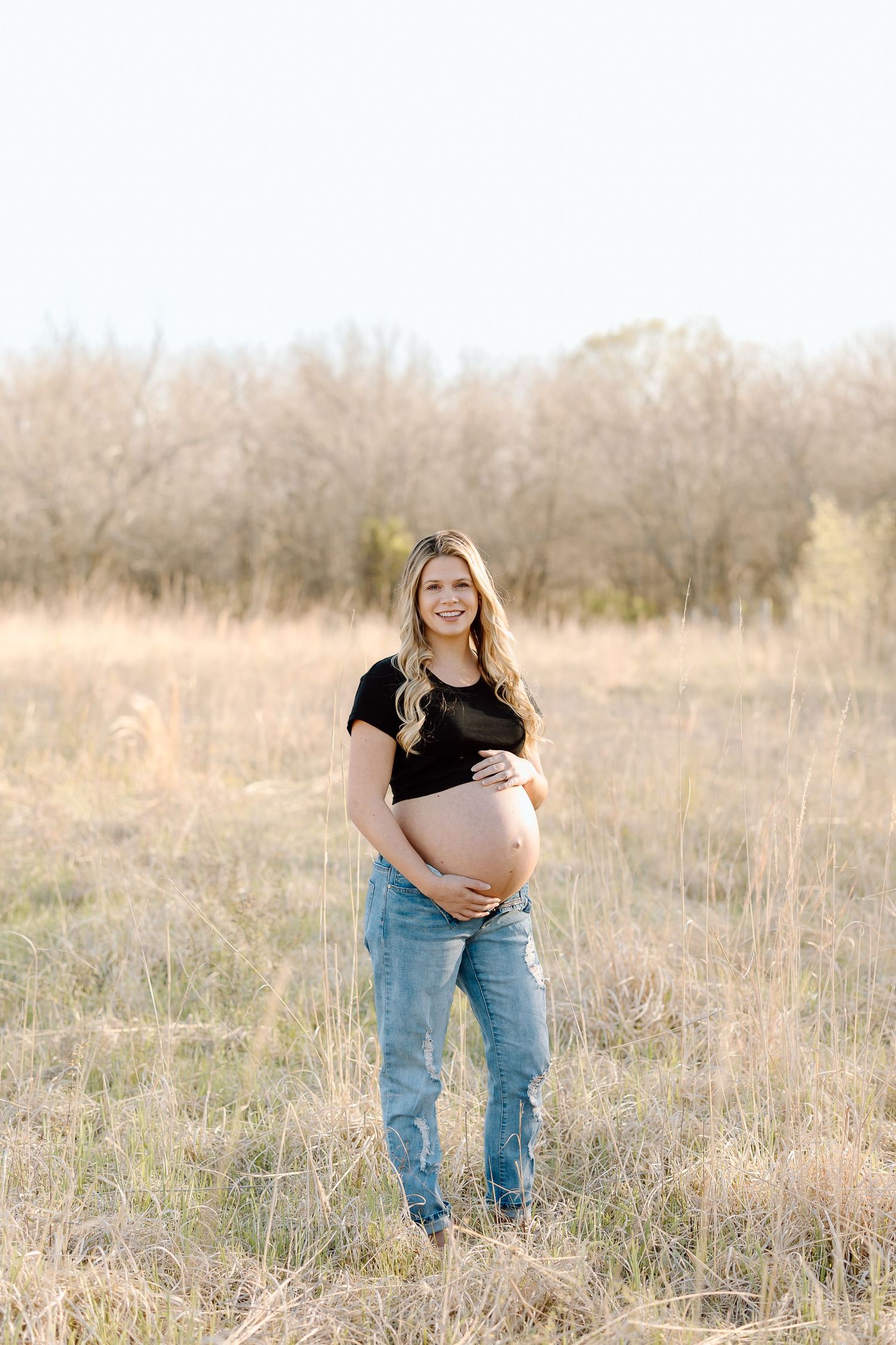 A pregnant woman in a black shirt and jeans stands in a golden field during sunset with trees in the background.