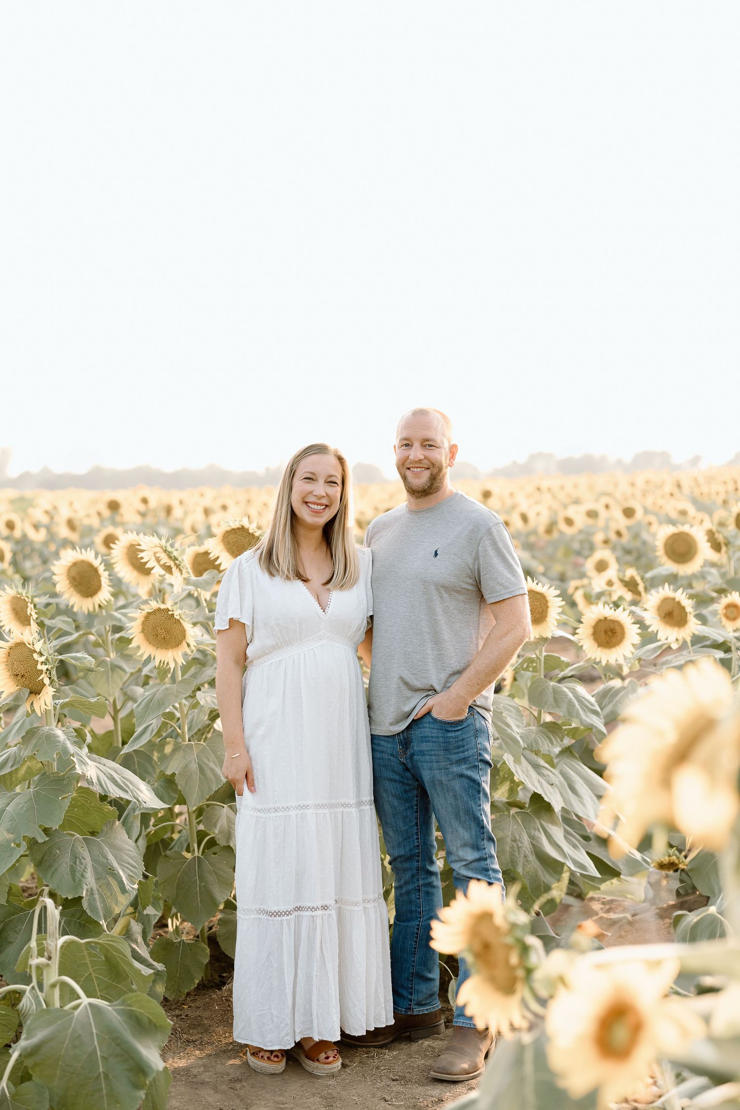 A couple stands together in a beautiful sunflower field during golden hour, surrounded by bright yellow blooms.