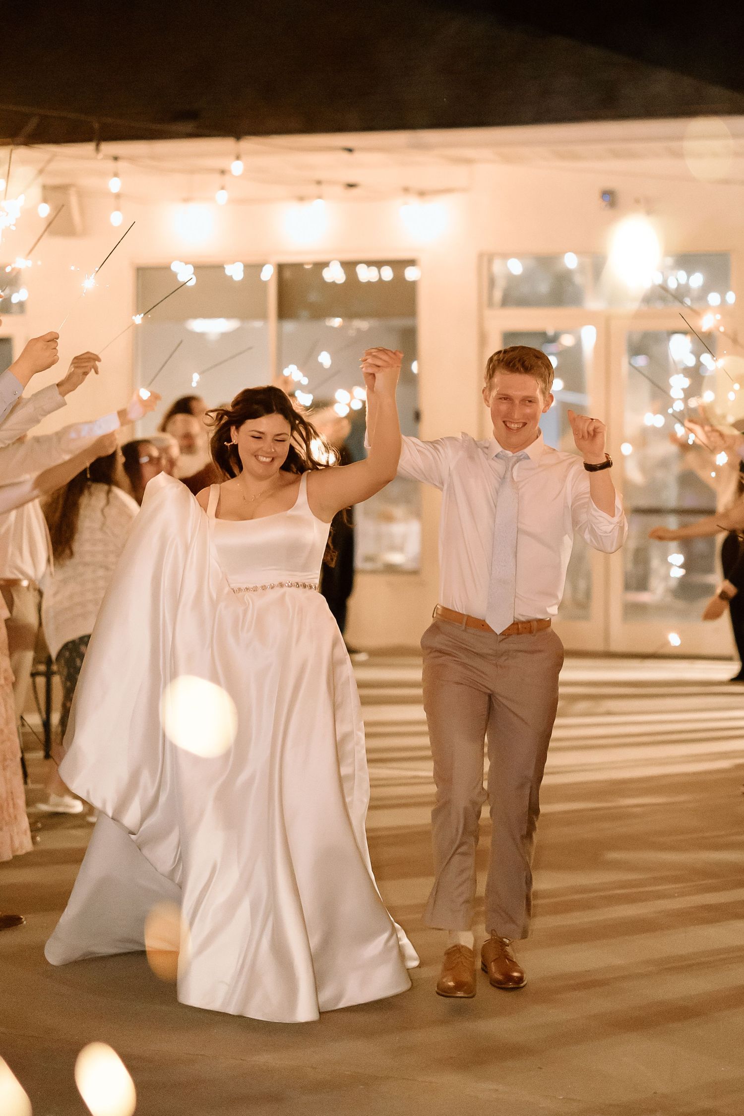 Couple in white wedding attire dances joyfully under twinkling lights in a parking garage.