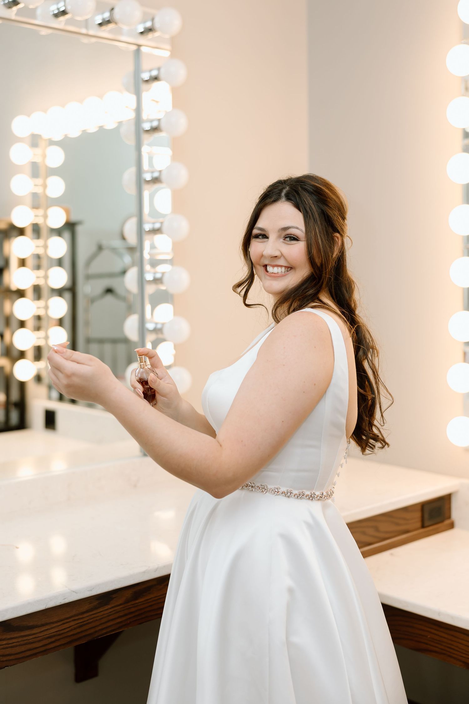 A bride in a white wedding dress smiles while standing in front of a lighted vanity mirror in a dressing room.