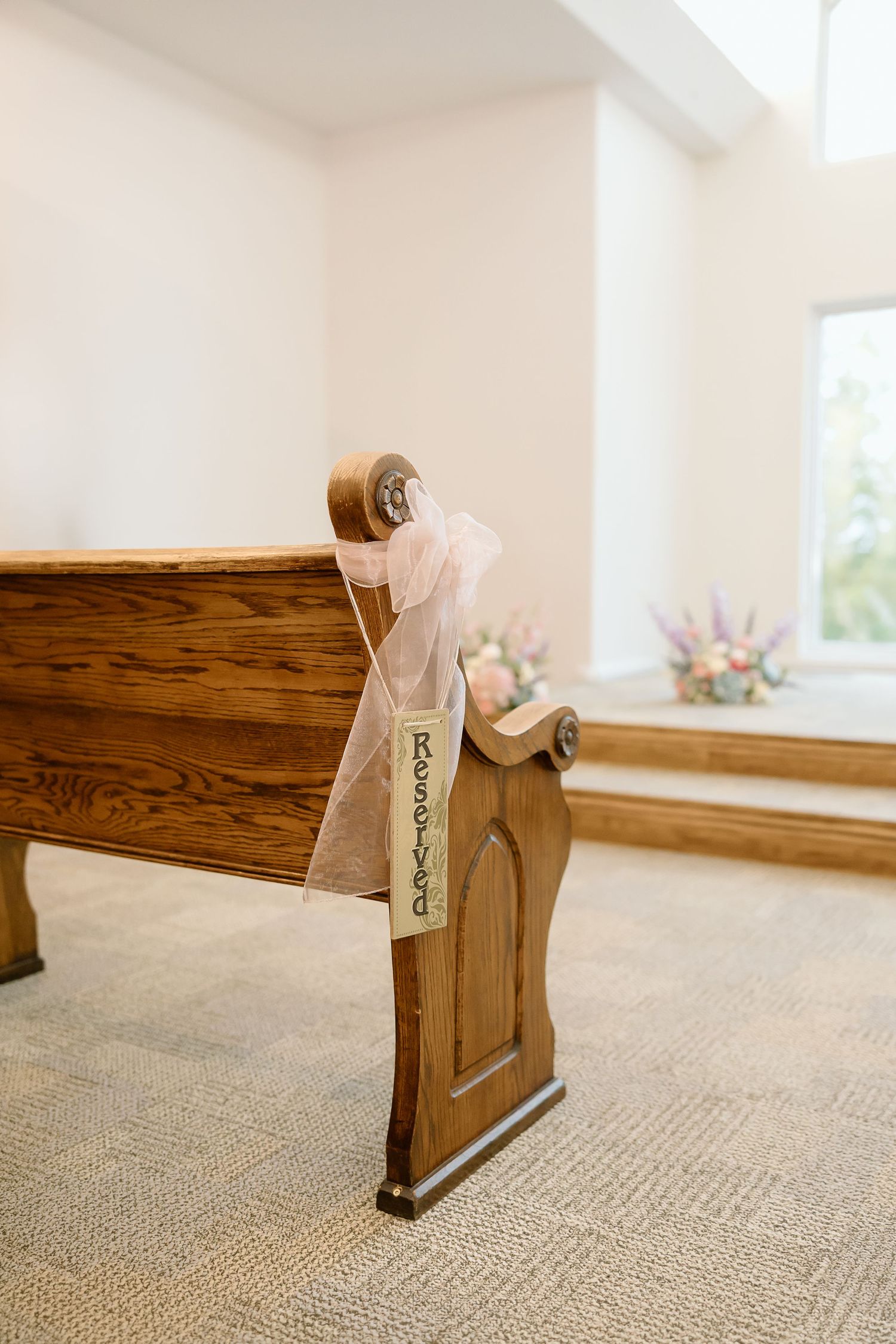 Wooden kneeler benches with beige cushions lined up in a row on carpet in a bright, sunlit church interior.