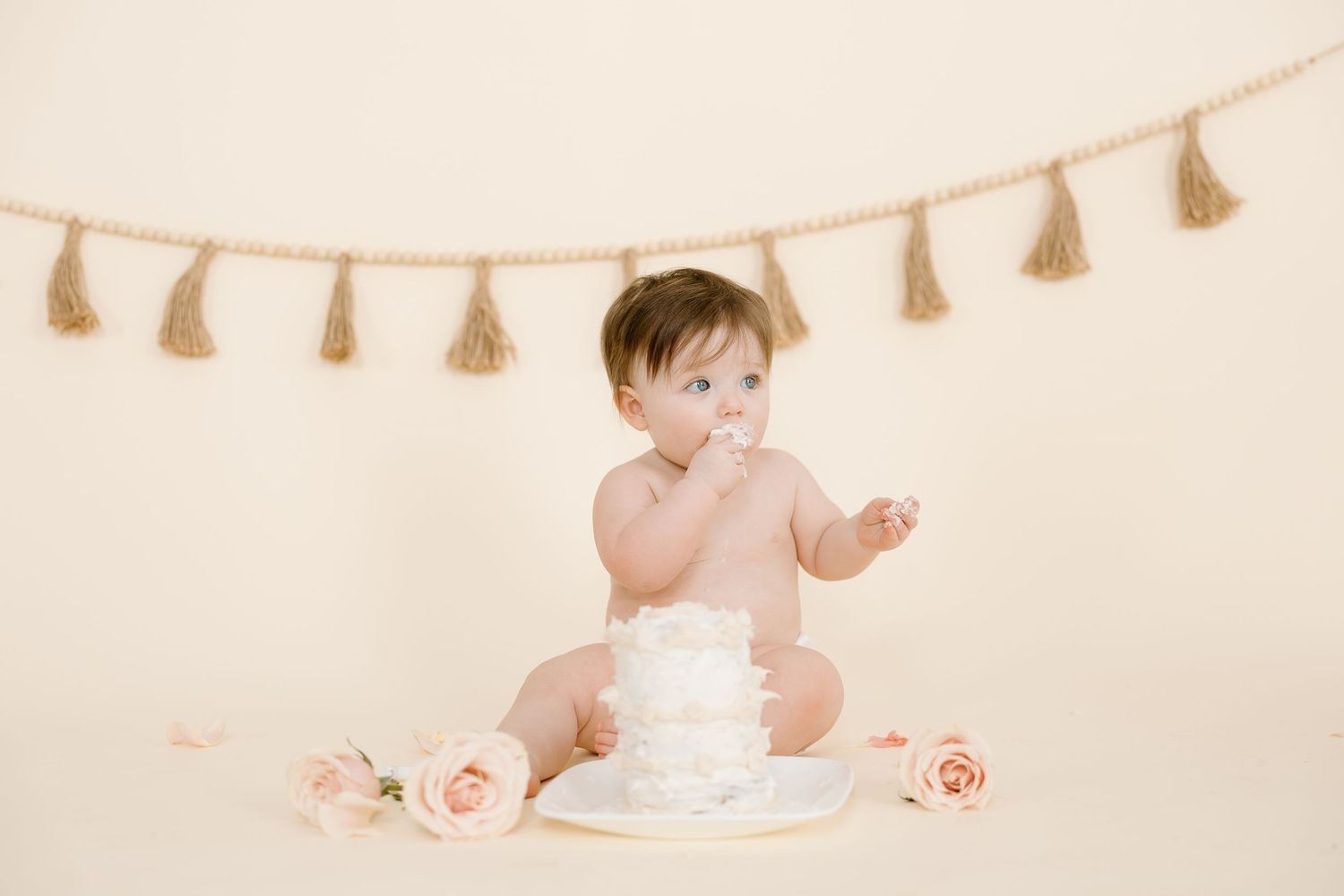 Baby explores white cake while sitting with pink roses beneath beige tassel garland decoration.