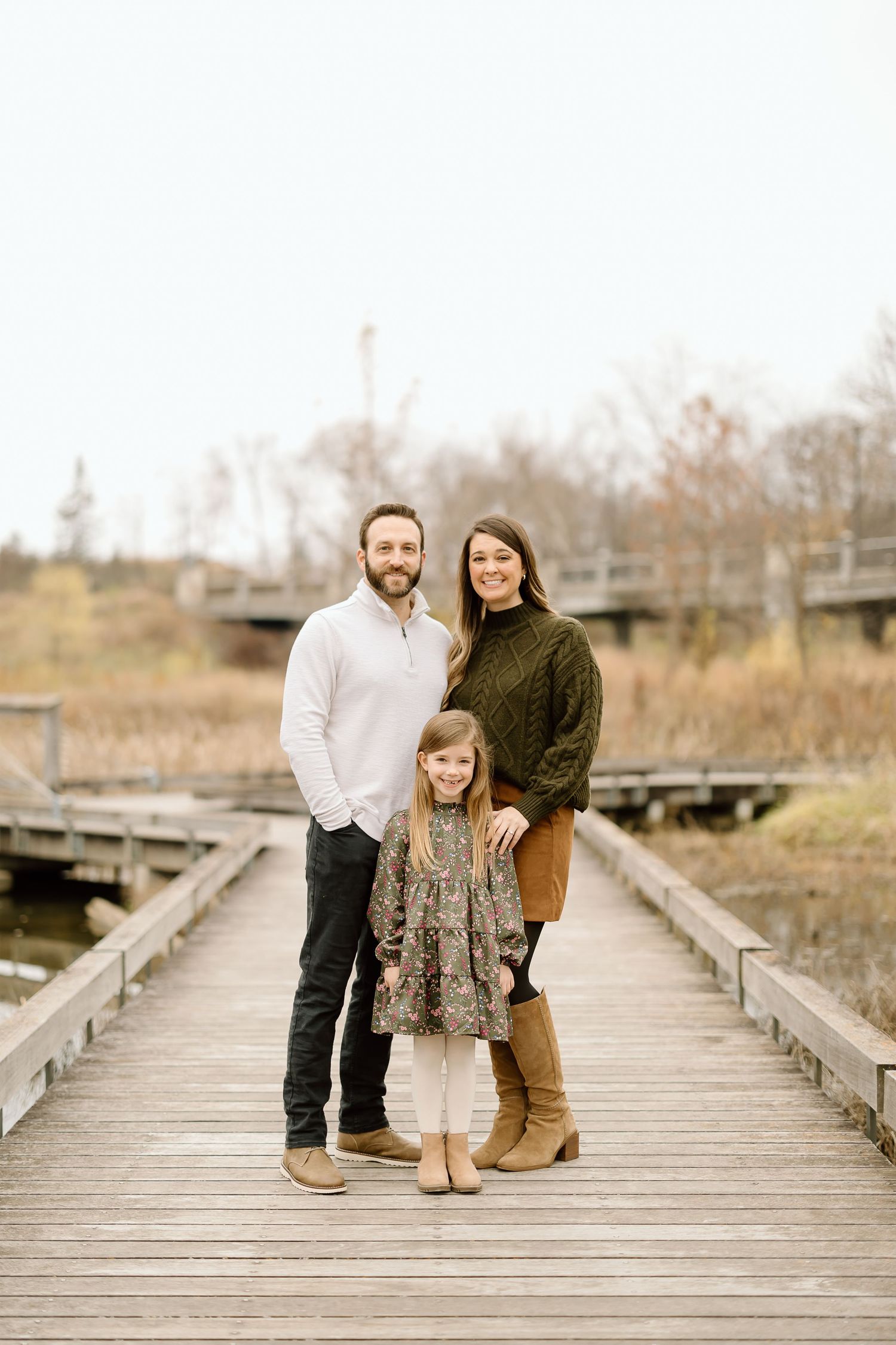Family of three poses together on wooden boardwalk in autumn landscape.