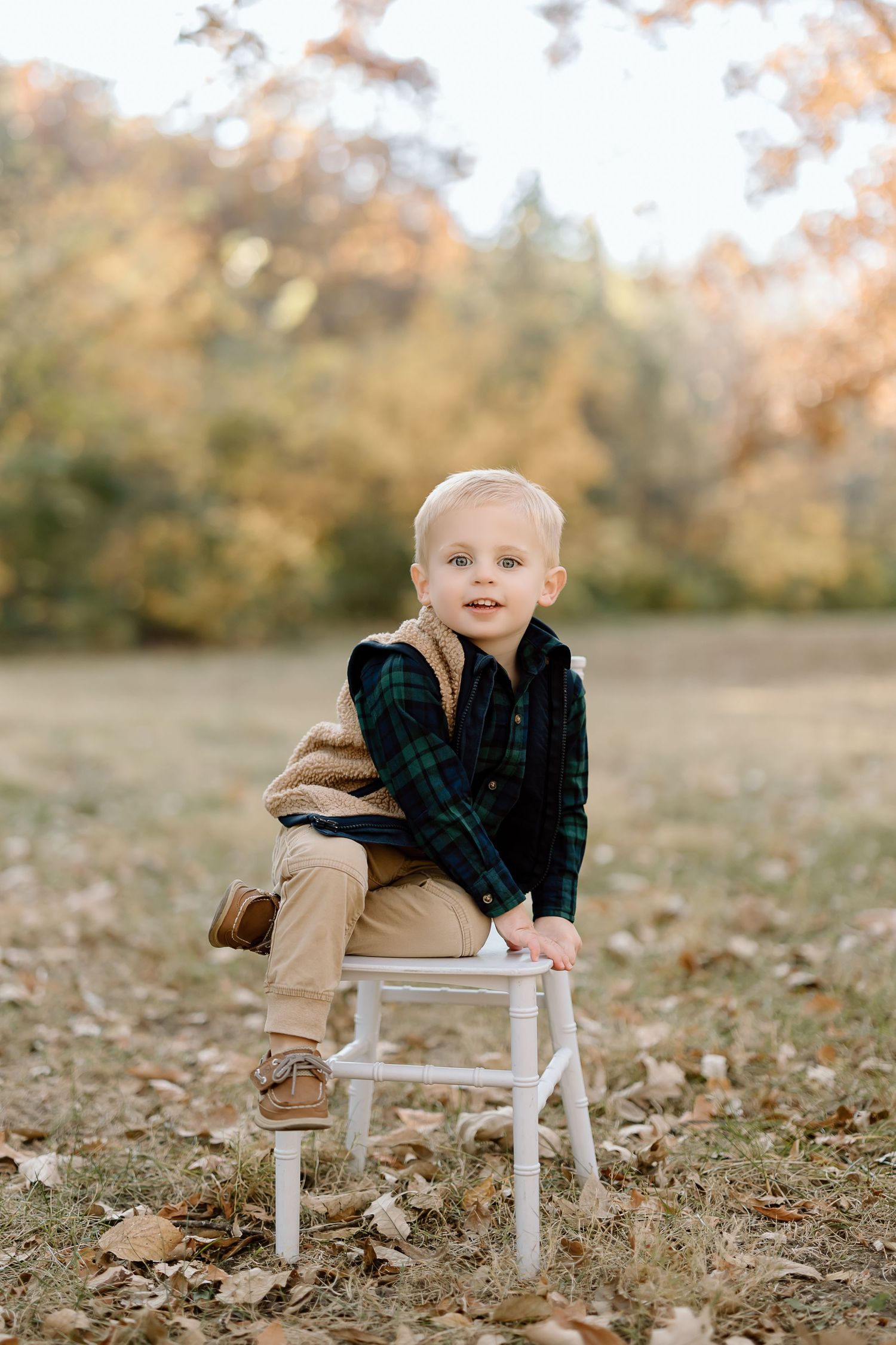Small child in plaid and vest sits on white stool among fallen autumn leaves.