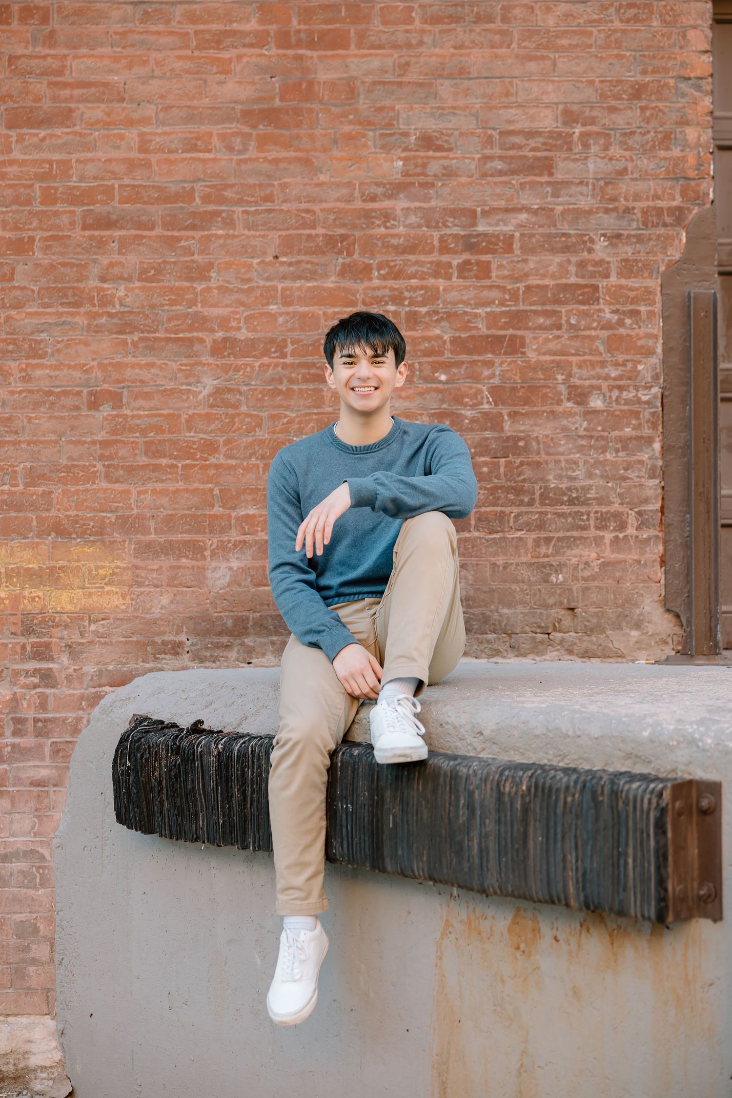 Person sitting on concrete ledge against brick wall in casual blue sweater.