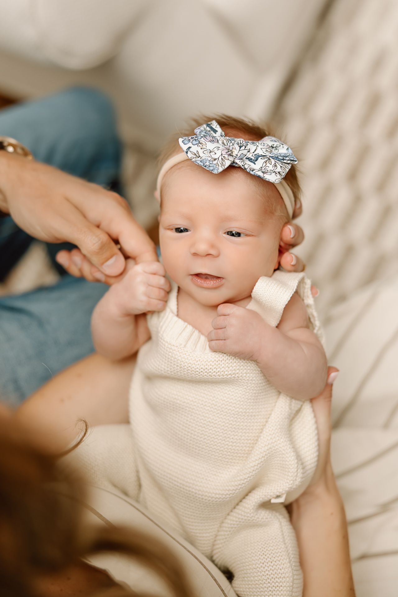 Baby wearing sparkly bow headband and cream knit outfit looking up with curious expression.