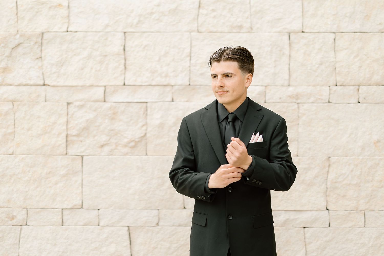 Groom in a classic black suit standing against a textured stone wall.