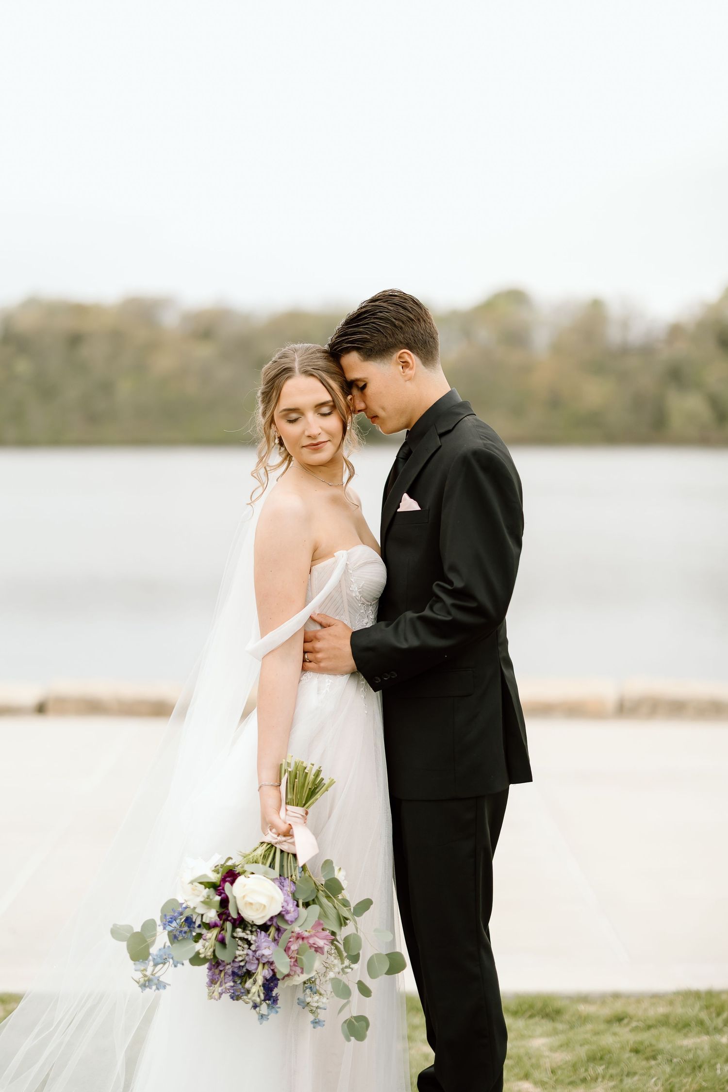 Elegant wedding couple shares tender moment while holding purple and white bouquet.