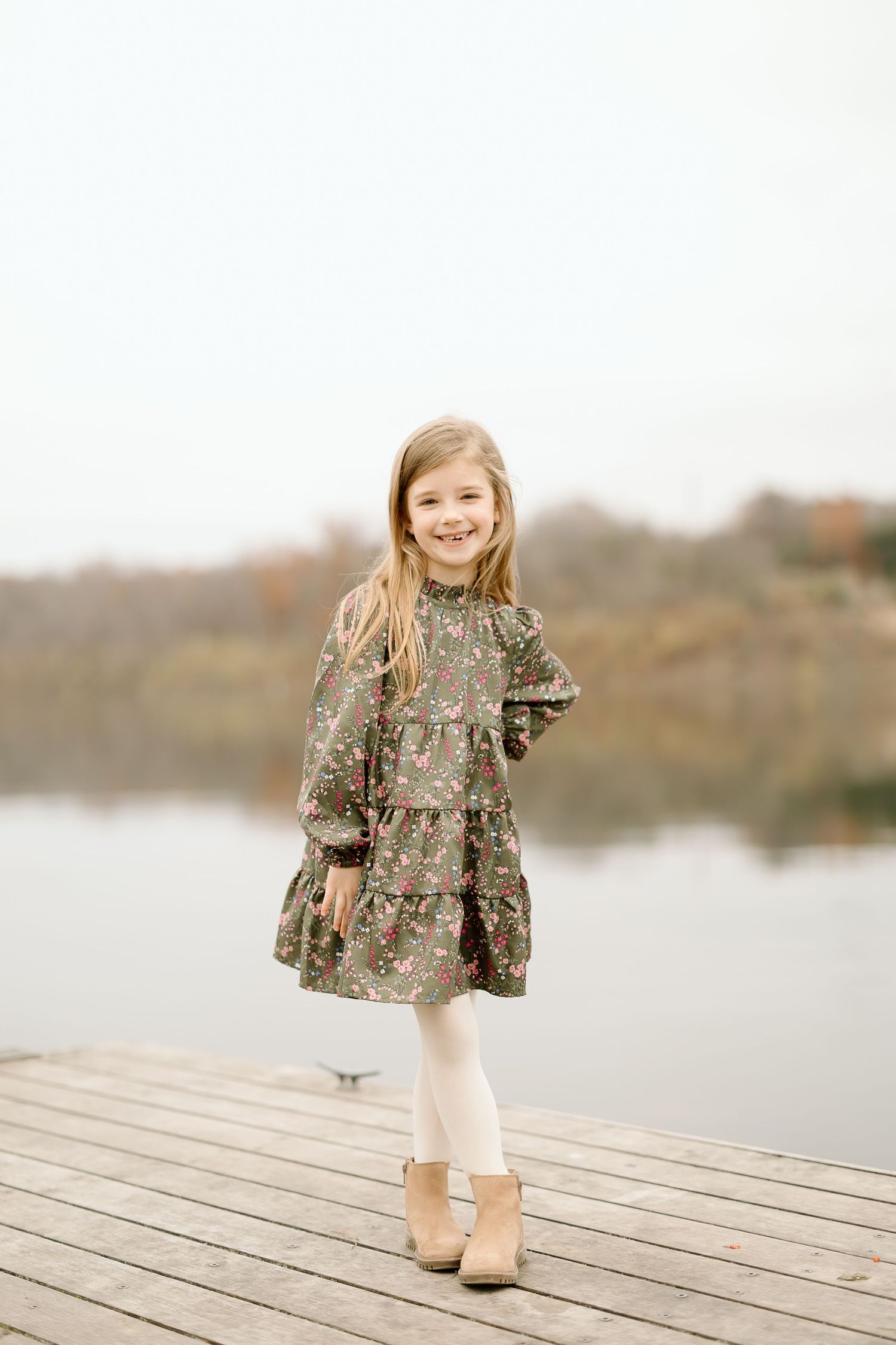 Young person in a patterned dress and boots posing on wooden boardwalk near water.