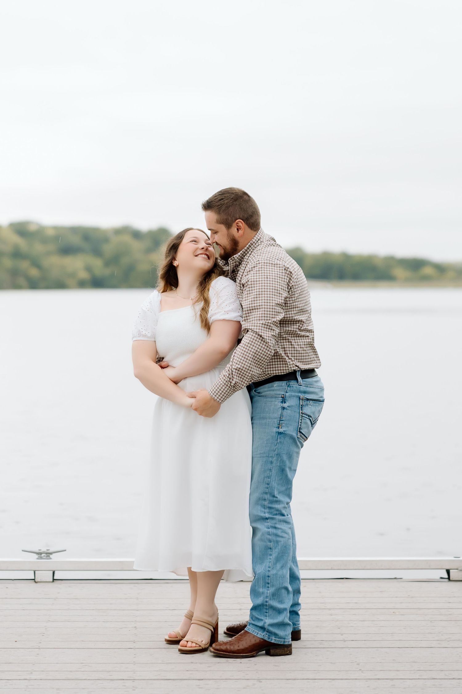 A couple shares a romantic moment in white and casual attire by a serene lake.