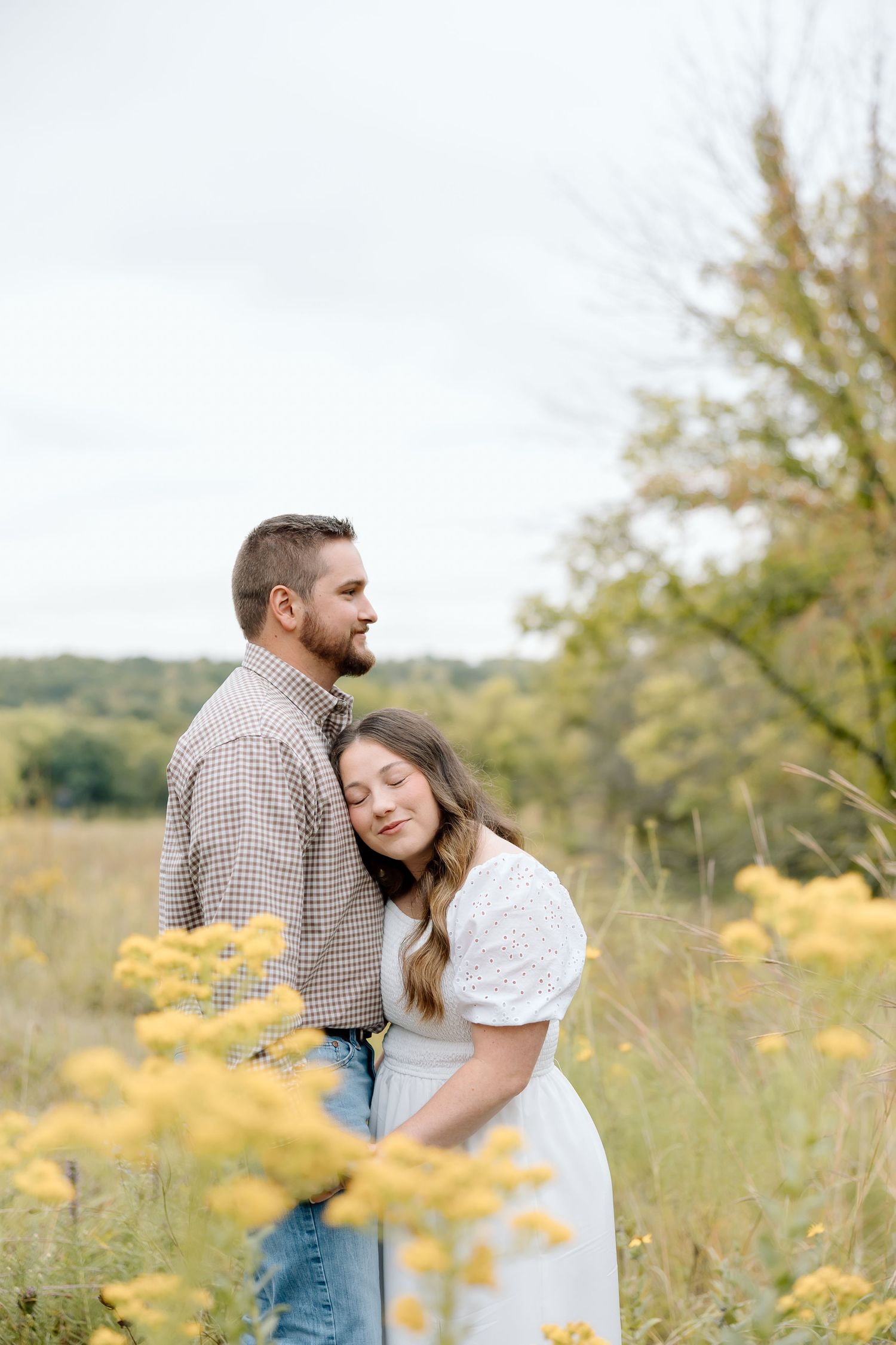 Couple shares gentle embrace while standing in field of yellow wildflowers on overcast day.