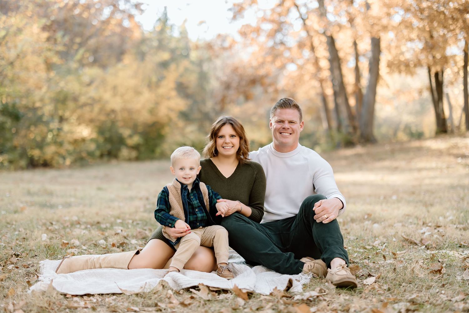 A family of three sits together on a blanket during an autumn photoshoot.