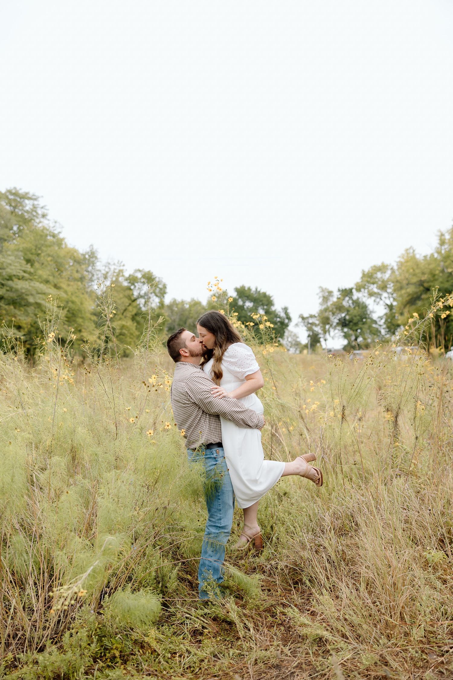 A couple shares a romantic lift pose in a sun-drenched meadow with tall grass and wildflowers.
