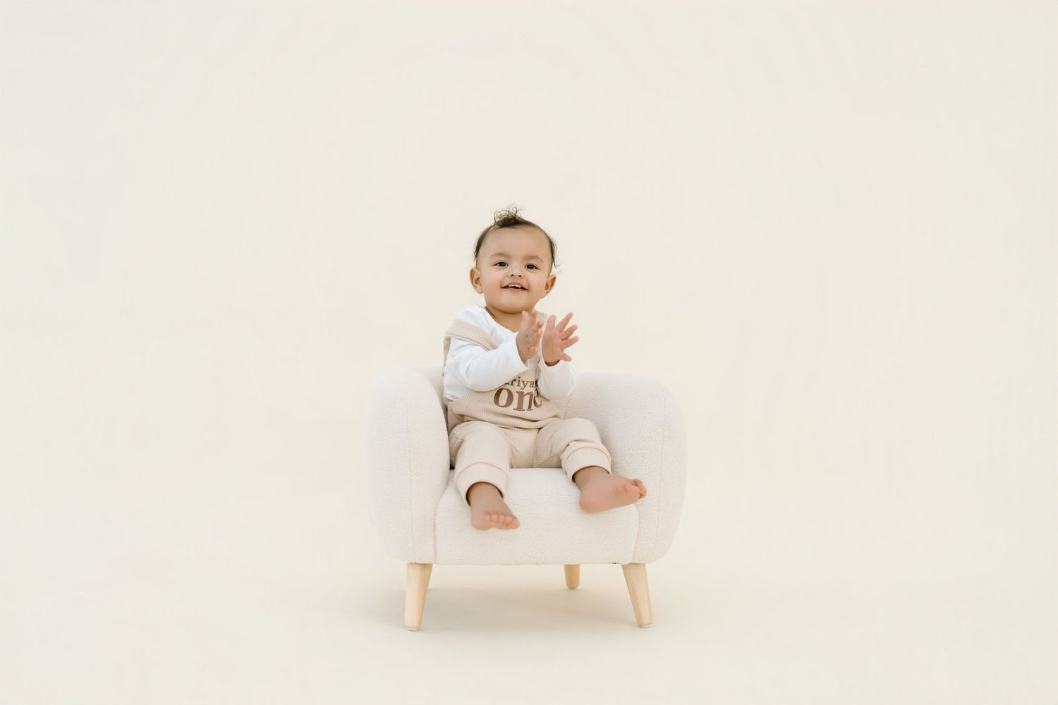 Baby wearing white onesie sits in small wooden chair against neutral background.