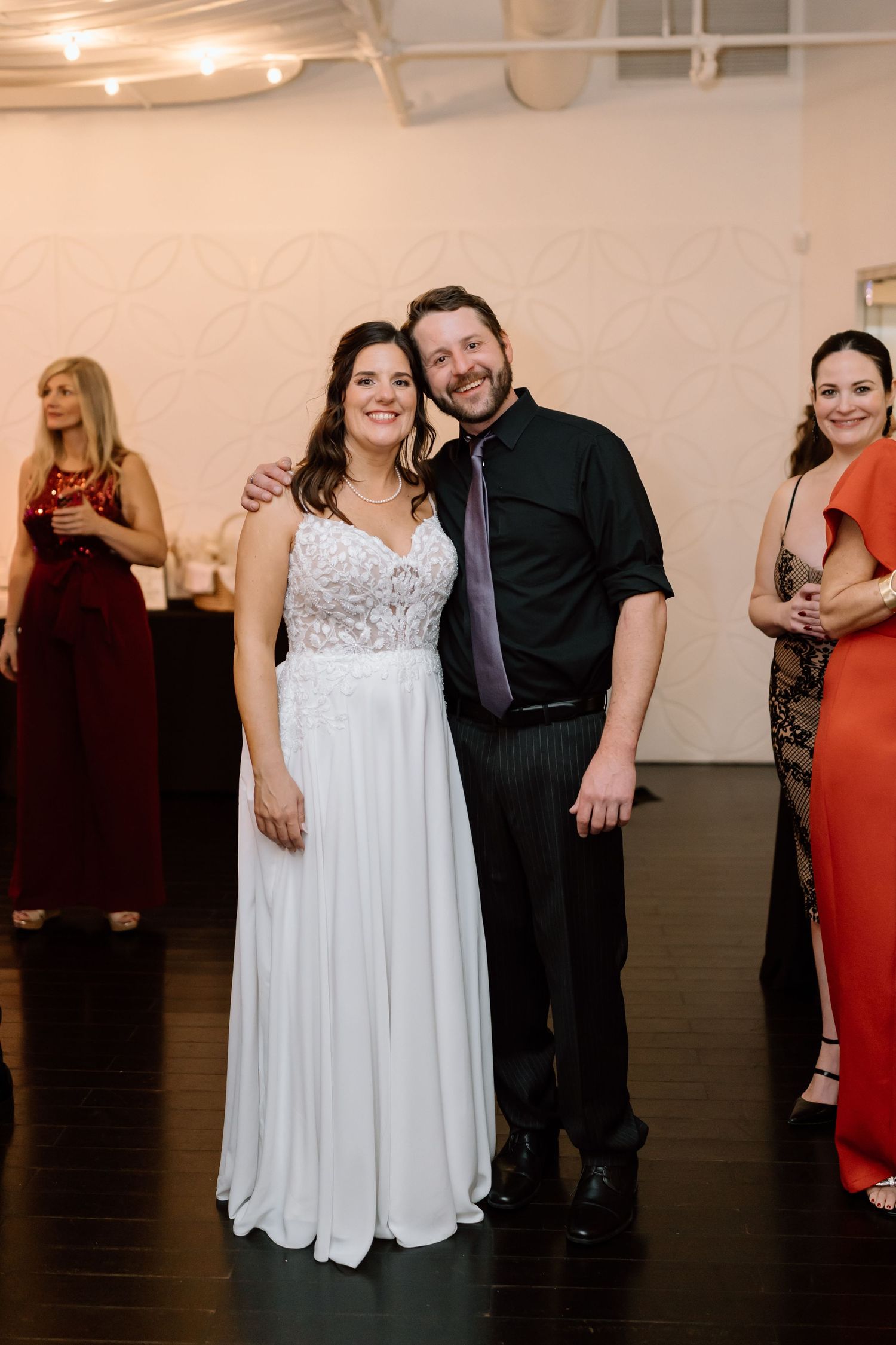 Couple posing at a formal event with guests in burgundy and coral dresses visible in the background.