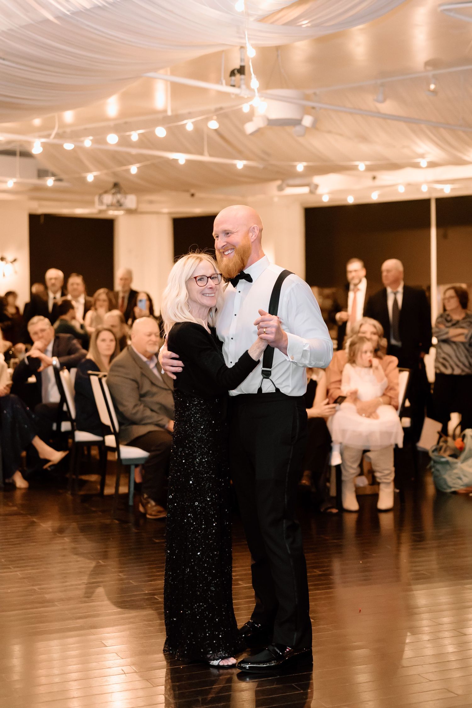 A couple shares an elegant dance moment on a wooden floor under draped ceiling with string lights at a formal event.