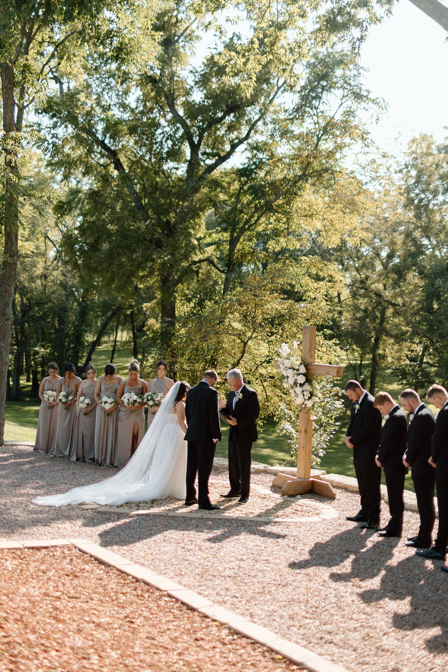 Wedding party stands in two lines facing each other on a gravel path surrounded by trees in an outdoor ceremony setting.