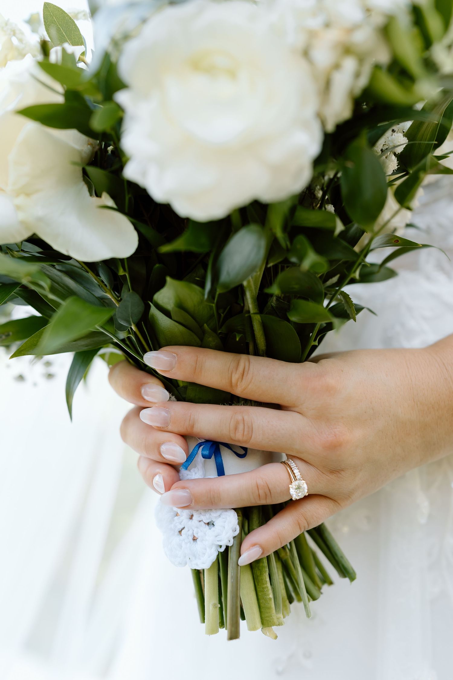 Close-up of hands holding elegant white peony bouquet with green foliage, showcasing engagement ring.