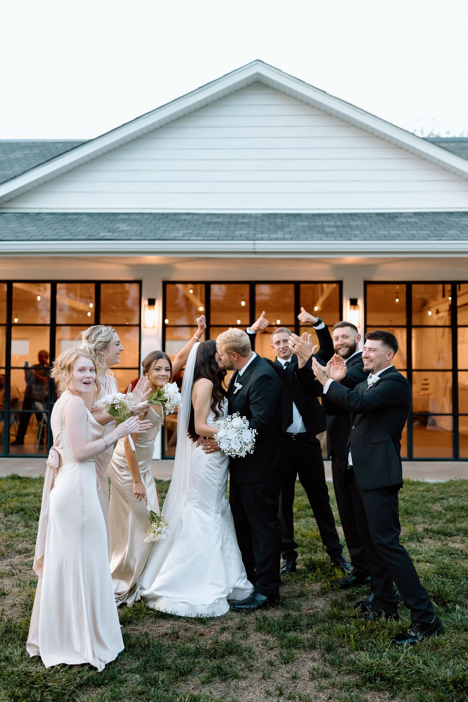 Wedding party celebrates with champagne toast outdoors near white building at sunset.
