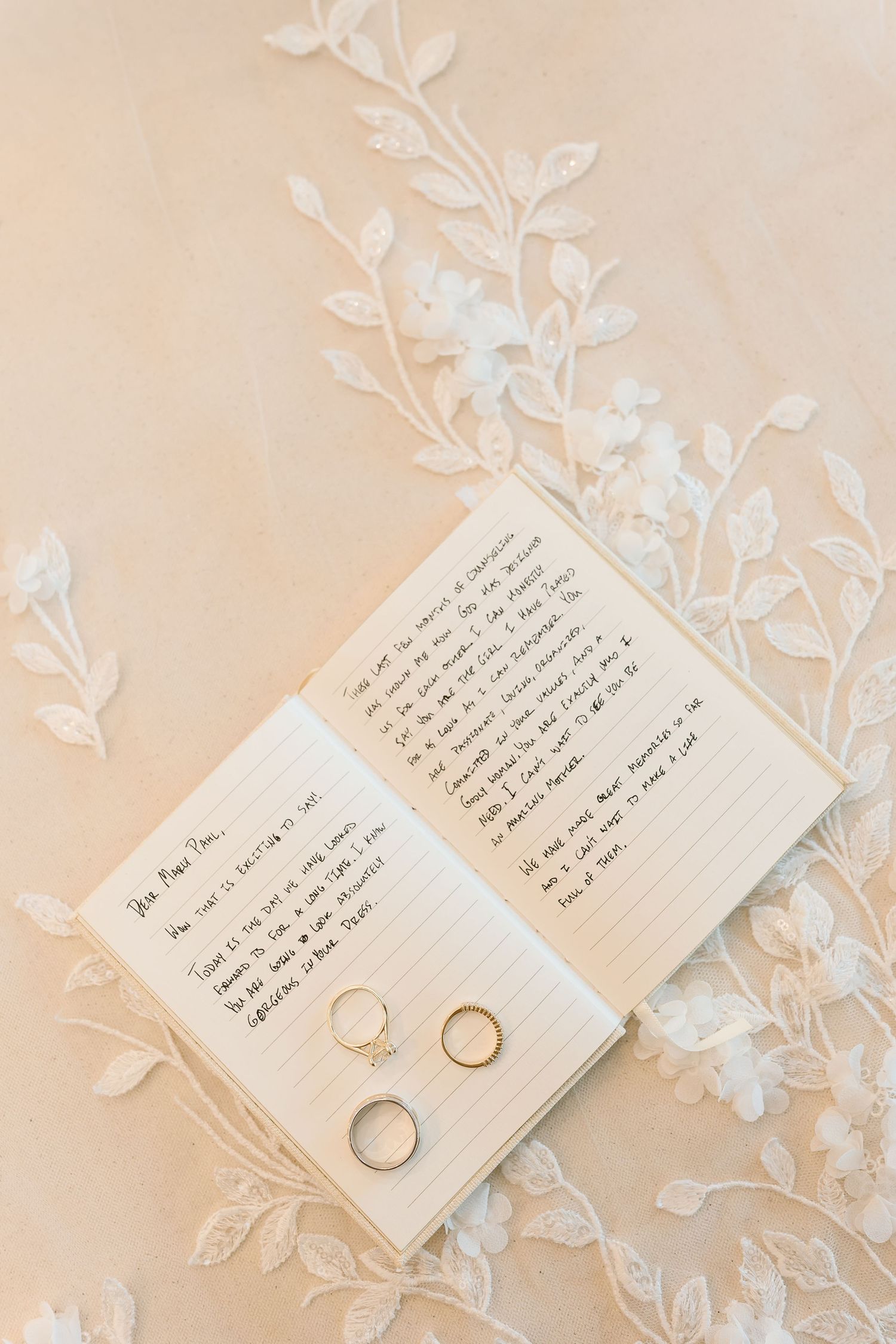 Wedding rings displayed on handwritten vows with delicate white floral decorations against a soft beige background.