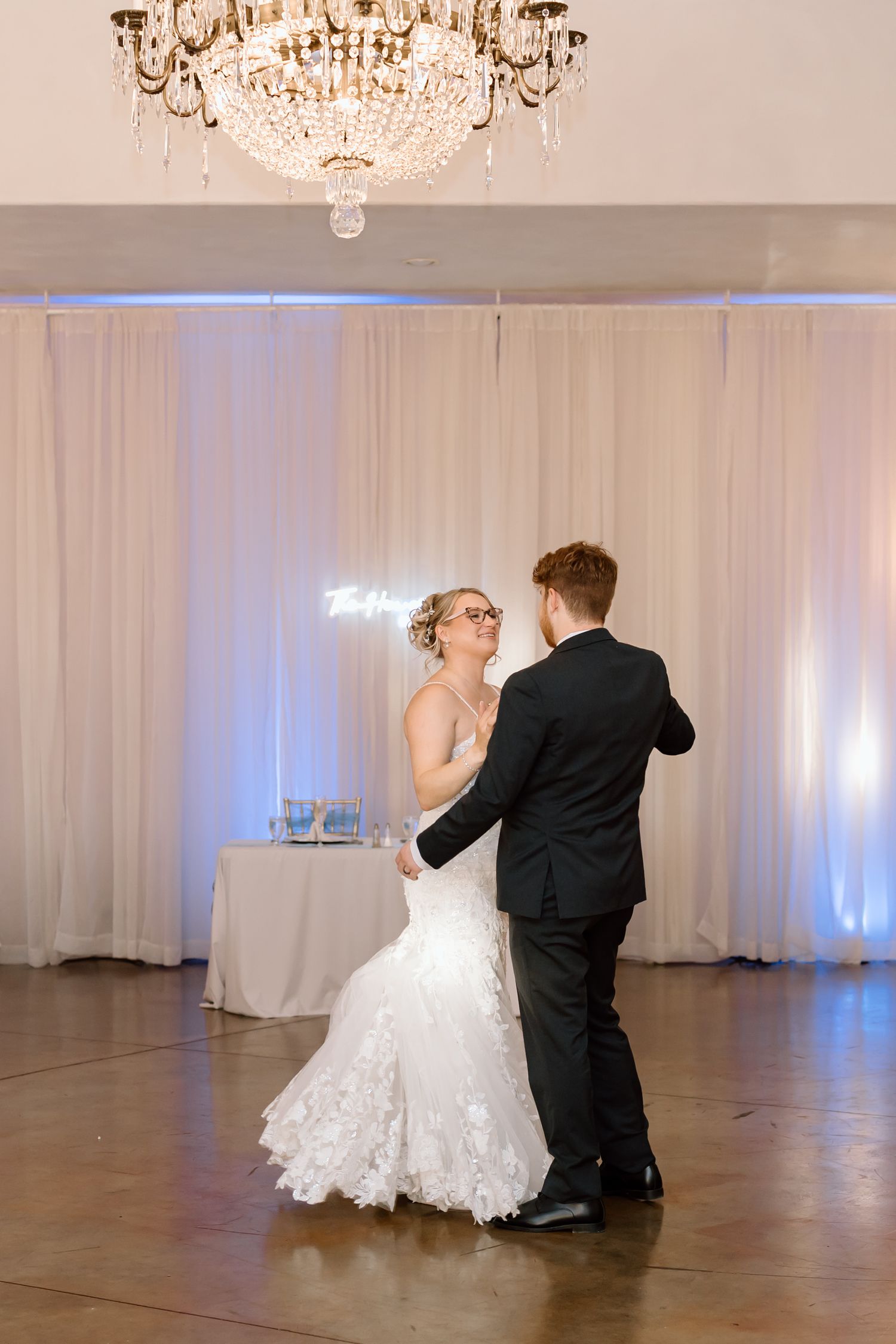 A couple in wedding attire performs a romantic dip during their first dance in an elegant ballroom with chandeliers.