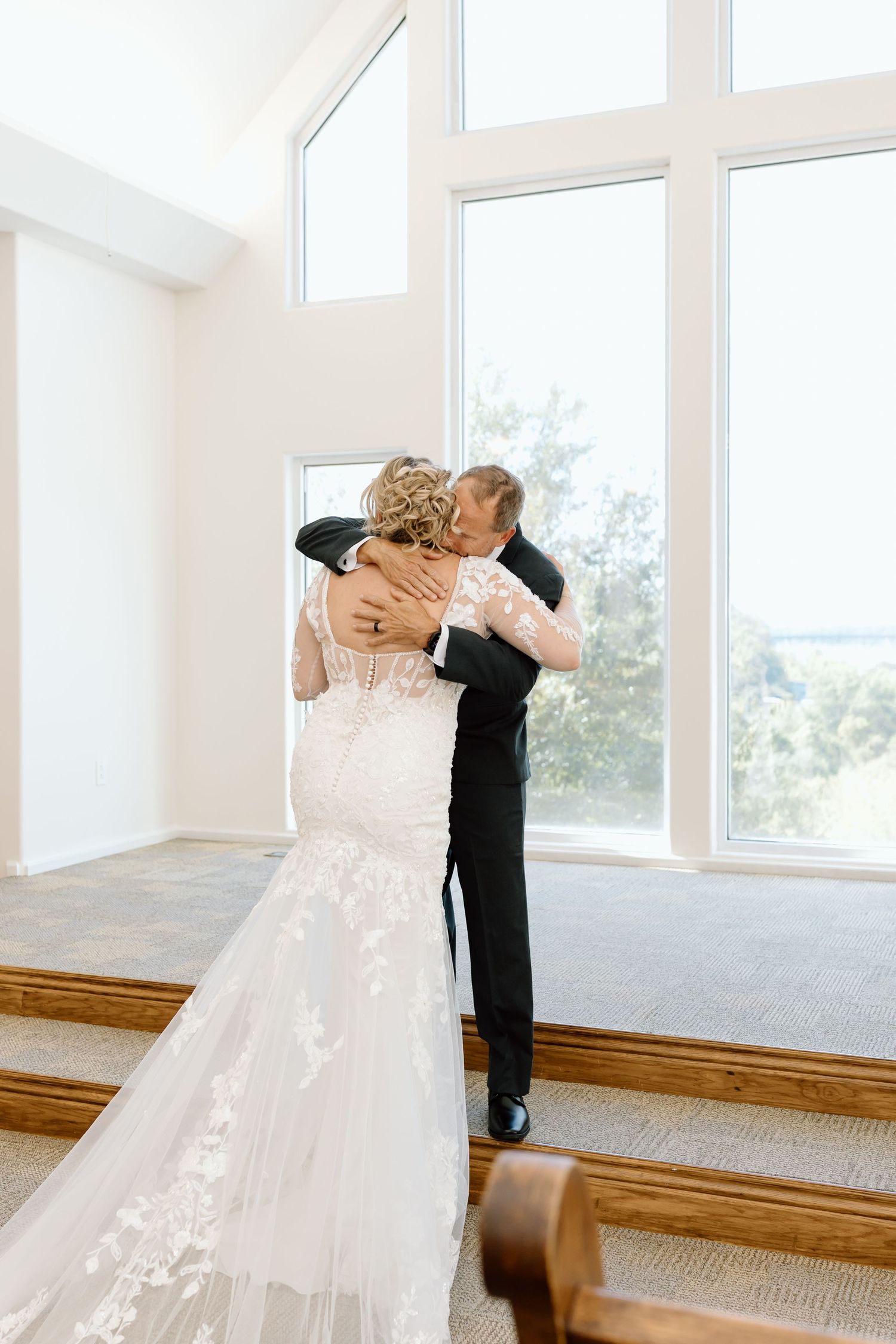 A couple in wedding attire share an intimate moment in a bright sunlit room with large windows.