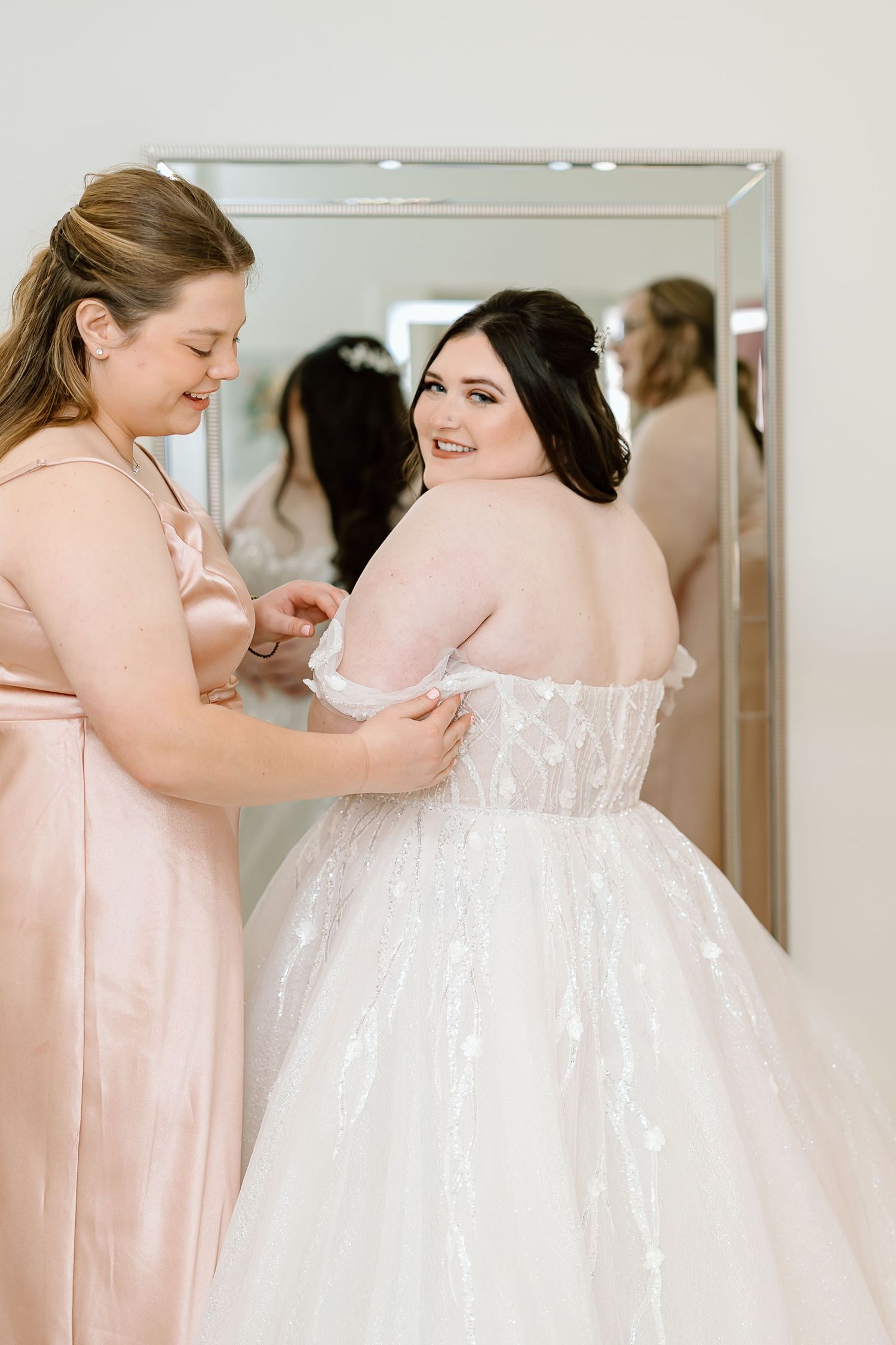A bridesmaid in pink helps adjust a white wedding gown during bridal preparations in a dressing room.