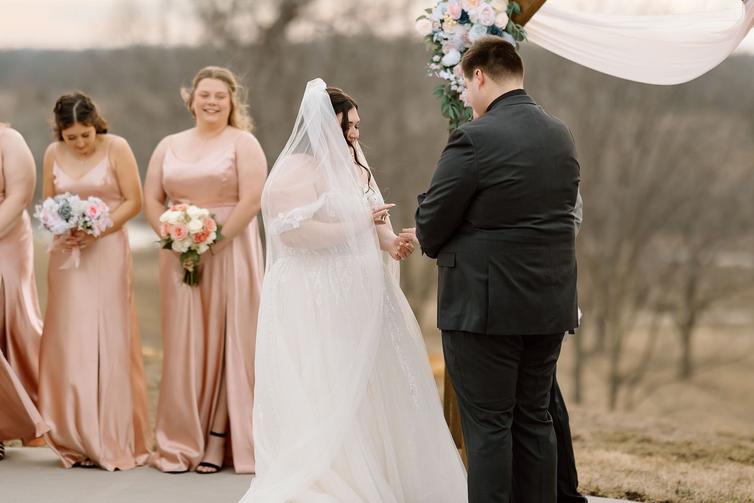 A wedding ceremony takes place outdoors with bridesmaids in blush pink dresses standing beside the couple under an arch.