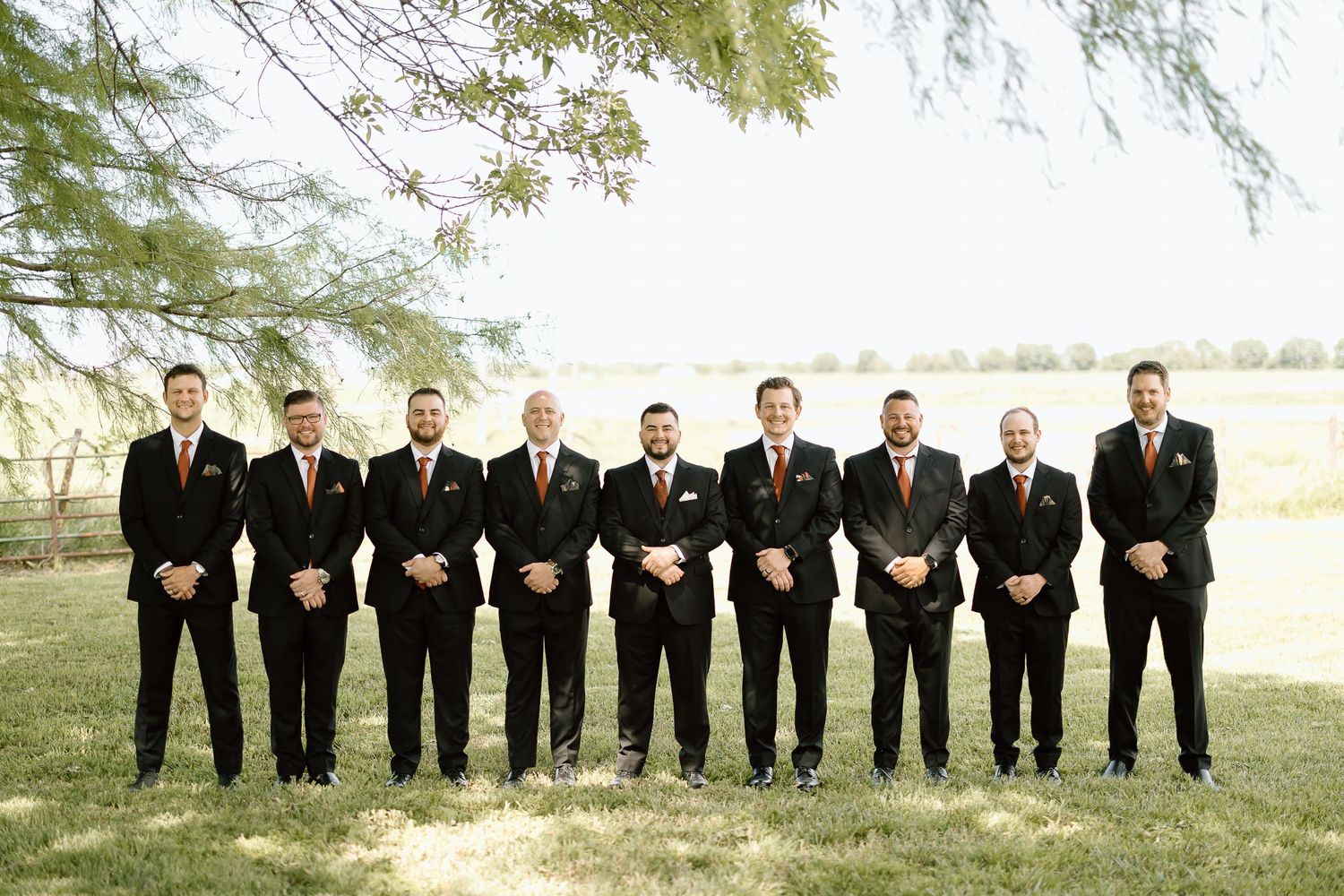 A row of groomsmen in black suits and ties pose together on a grassy slope for a wedding photo sequence.