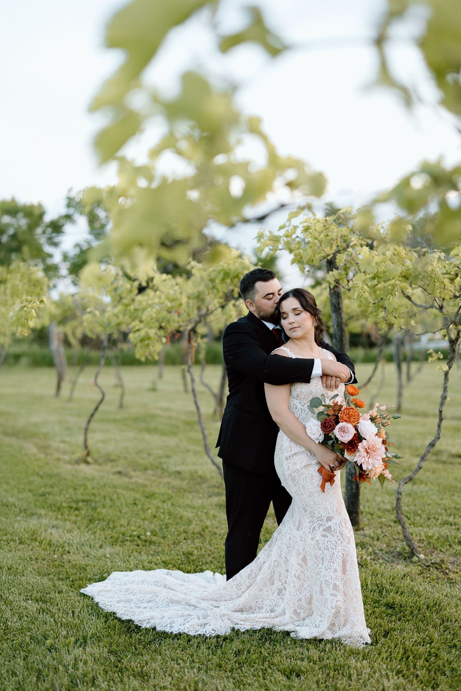 A couple in wedding attire embraces in a scenic orchard setting with the sun peeking through fruit trees.