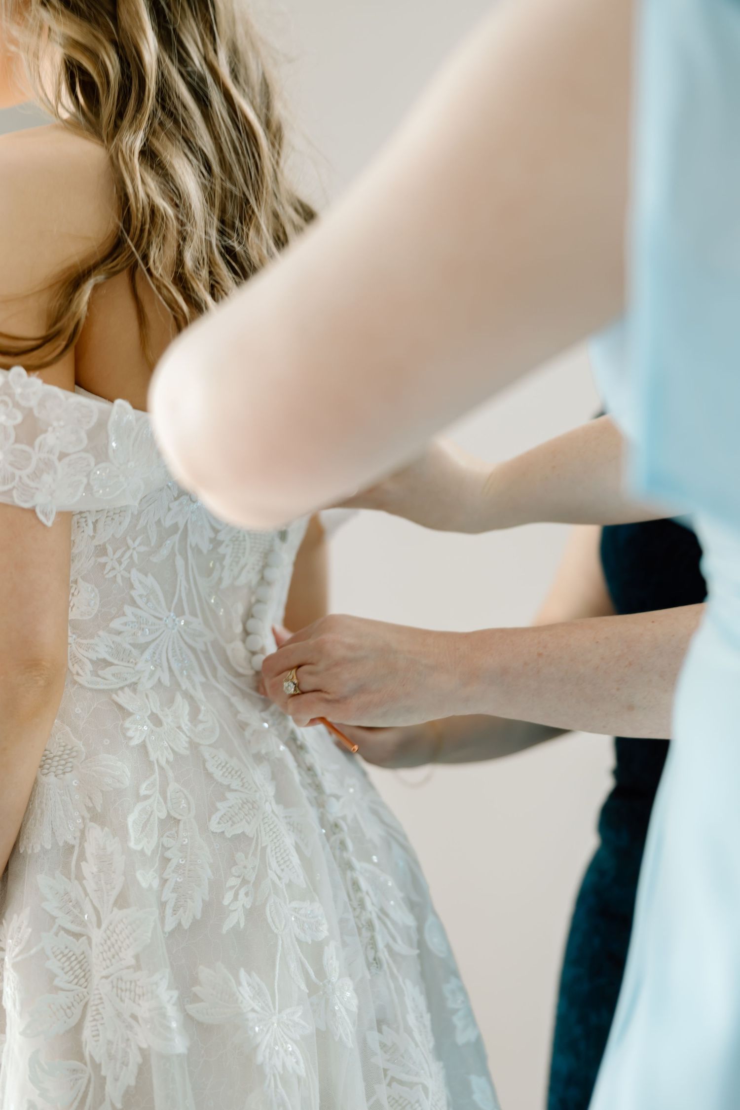 A bride in a white lace wedding gown gets her dress fastened by a helper, showcasing intricate floral lace details.