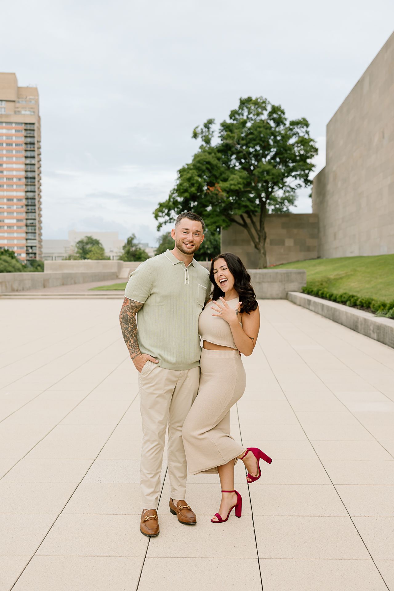 A couple in light beige outfits playfully poses together on concrete steps with red heels adding a pop of color.