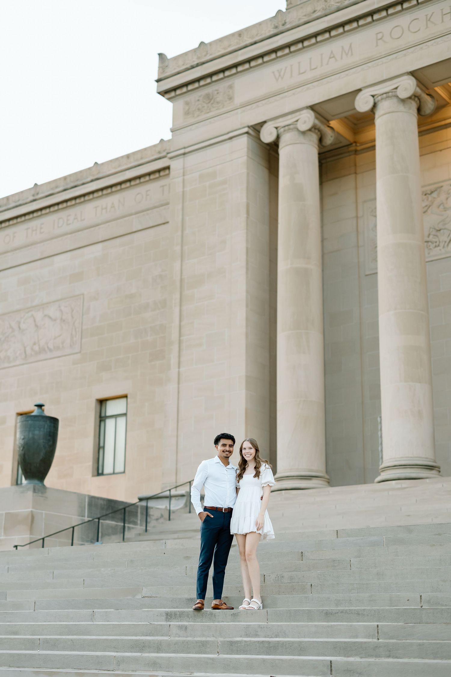 A couple stands on the grand stone steps of a neoclassical building with tall columns and ornate architecture.