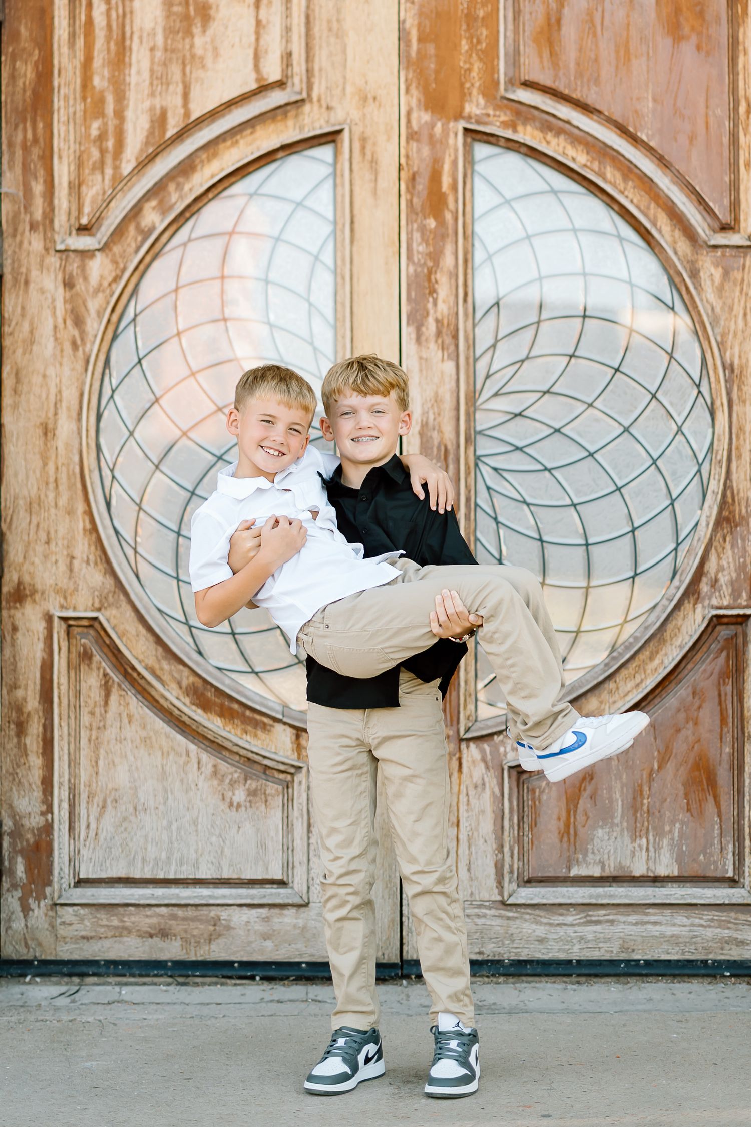 Two young children in matching outfits play and hug in front of a rustic wooden door with circular windows.