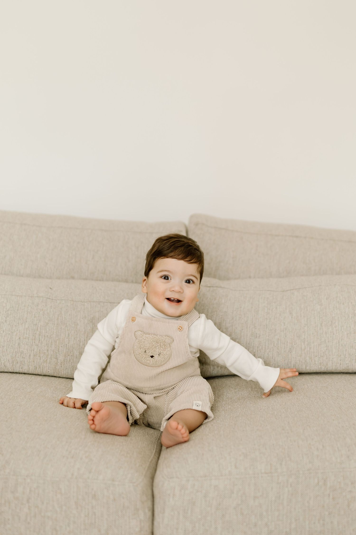 Baby in neutral-colored clothing sits on a beige couch in a minimalist setting, showing a progression of poses.