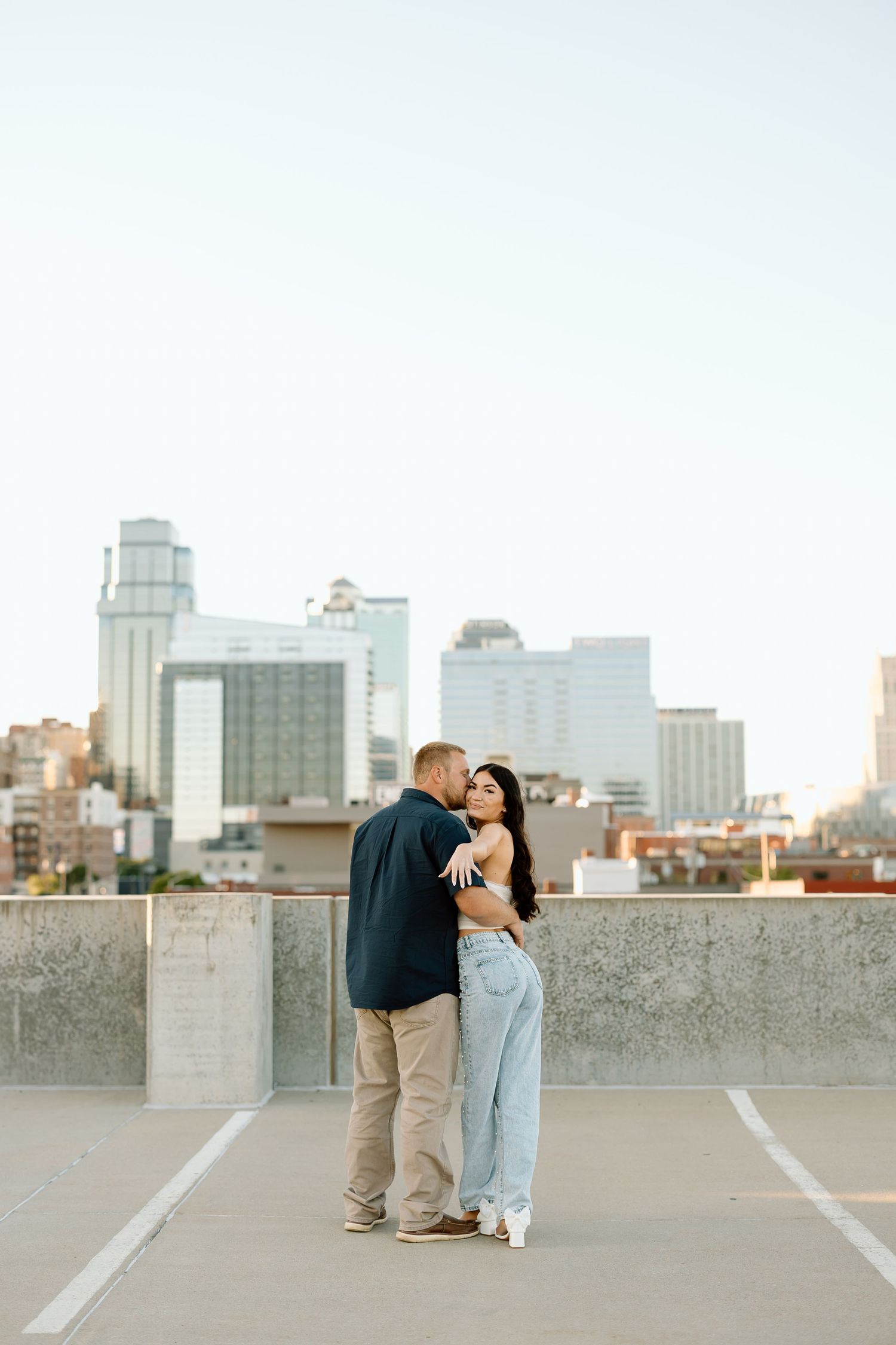 A couple shares a romantic moment on an urban rooftop parking area with city buildings visible in the background.