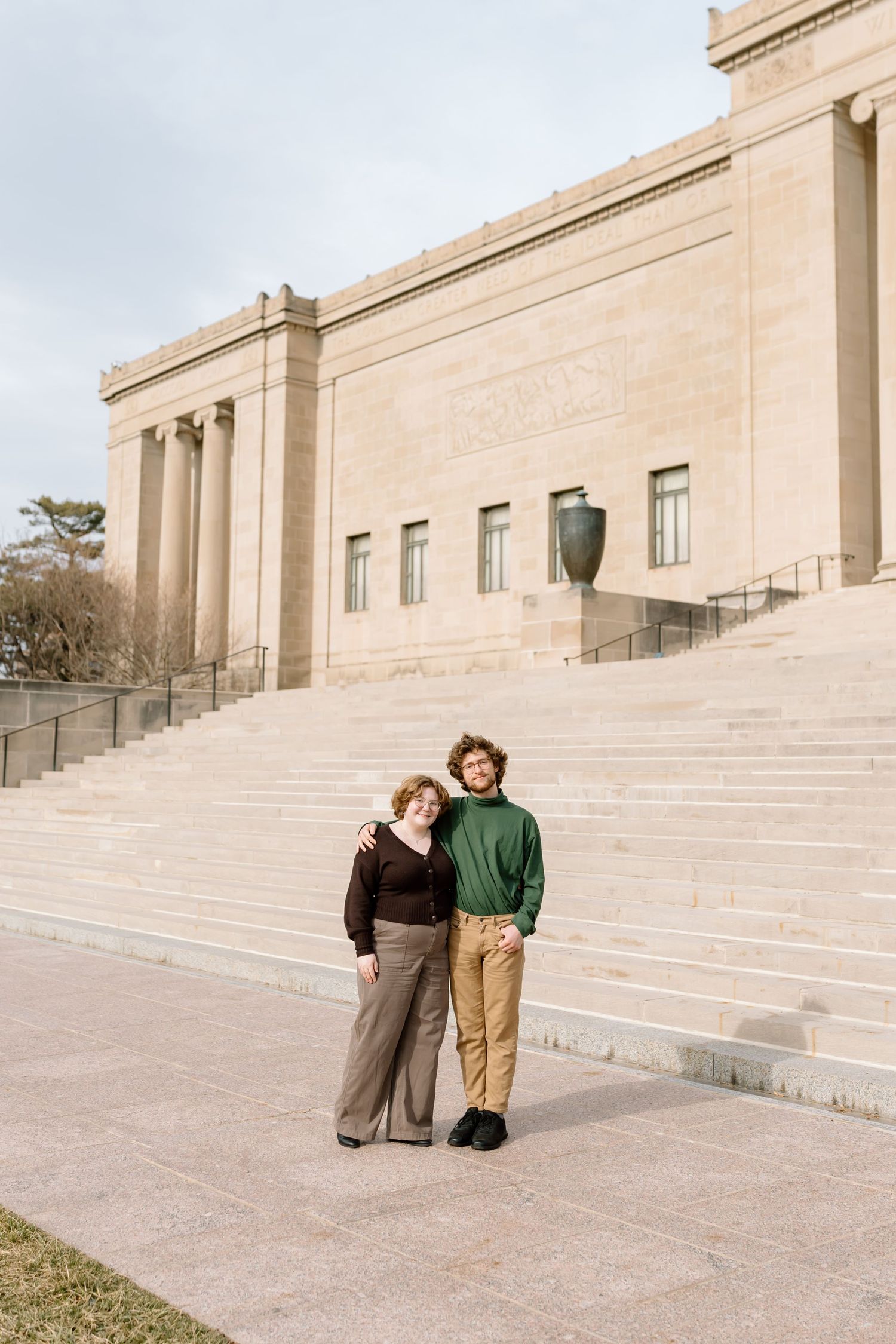 Two people stand together on wide stone steps of a grand neoclassical building with beige walls.