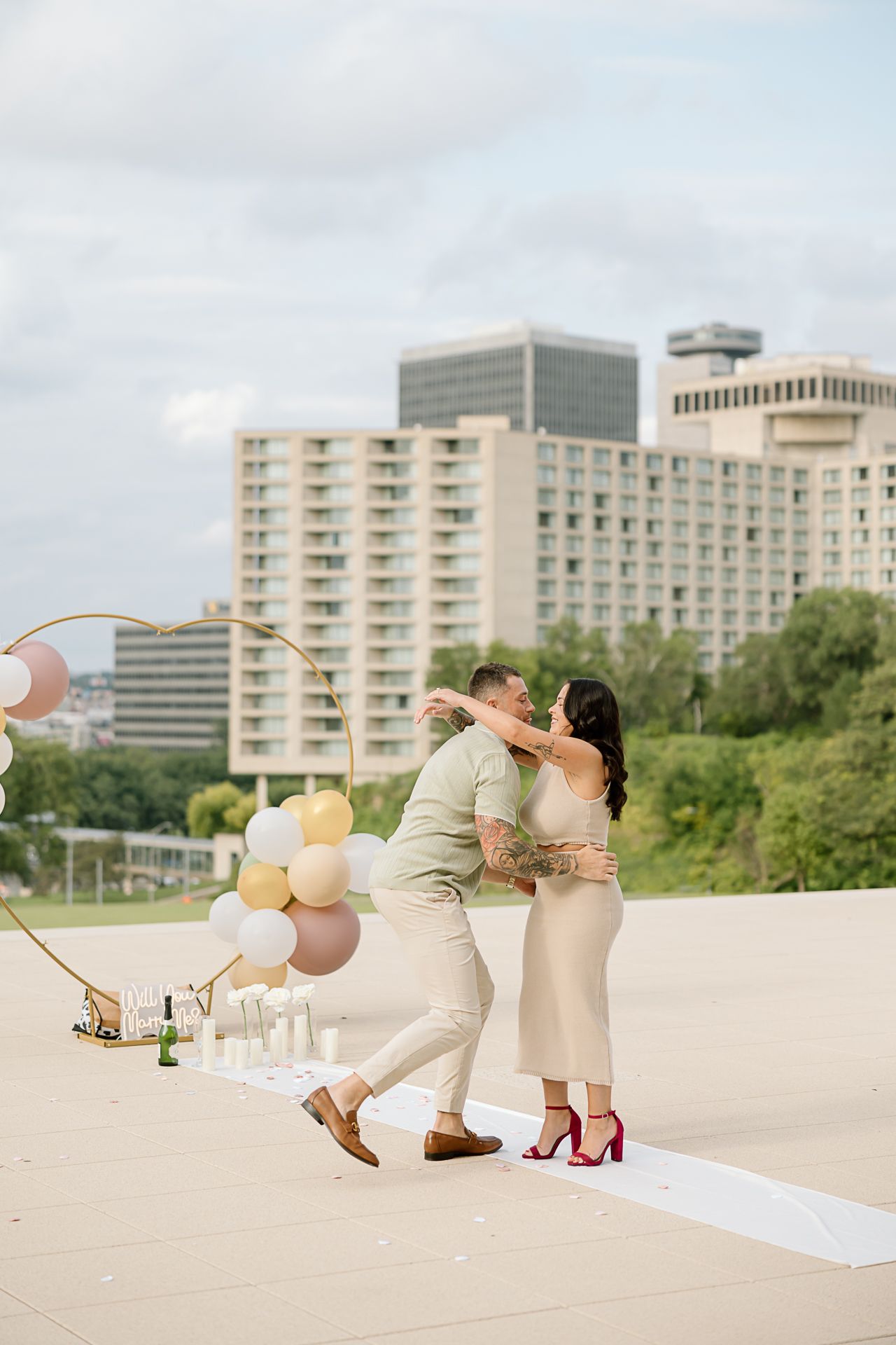 A couple shares a romantic moment on a rooftop with pastel balloons and city buildings in the background.