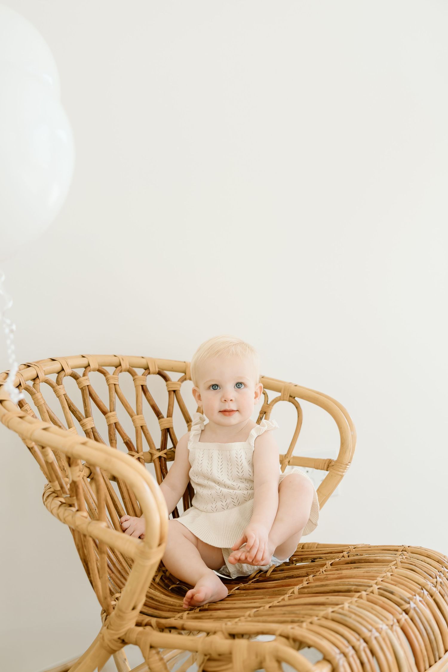 Small baby wearing cream sweater sits in vintage rattan chair against white background.