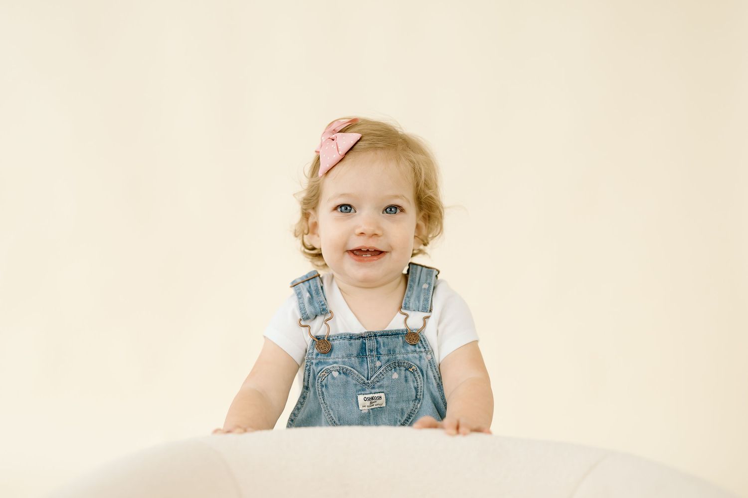 A child in denim overalls smiles while sitting against a light beige background.