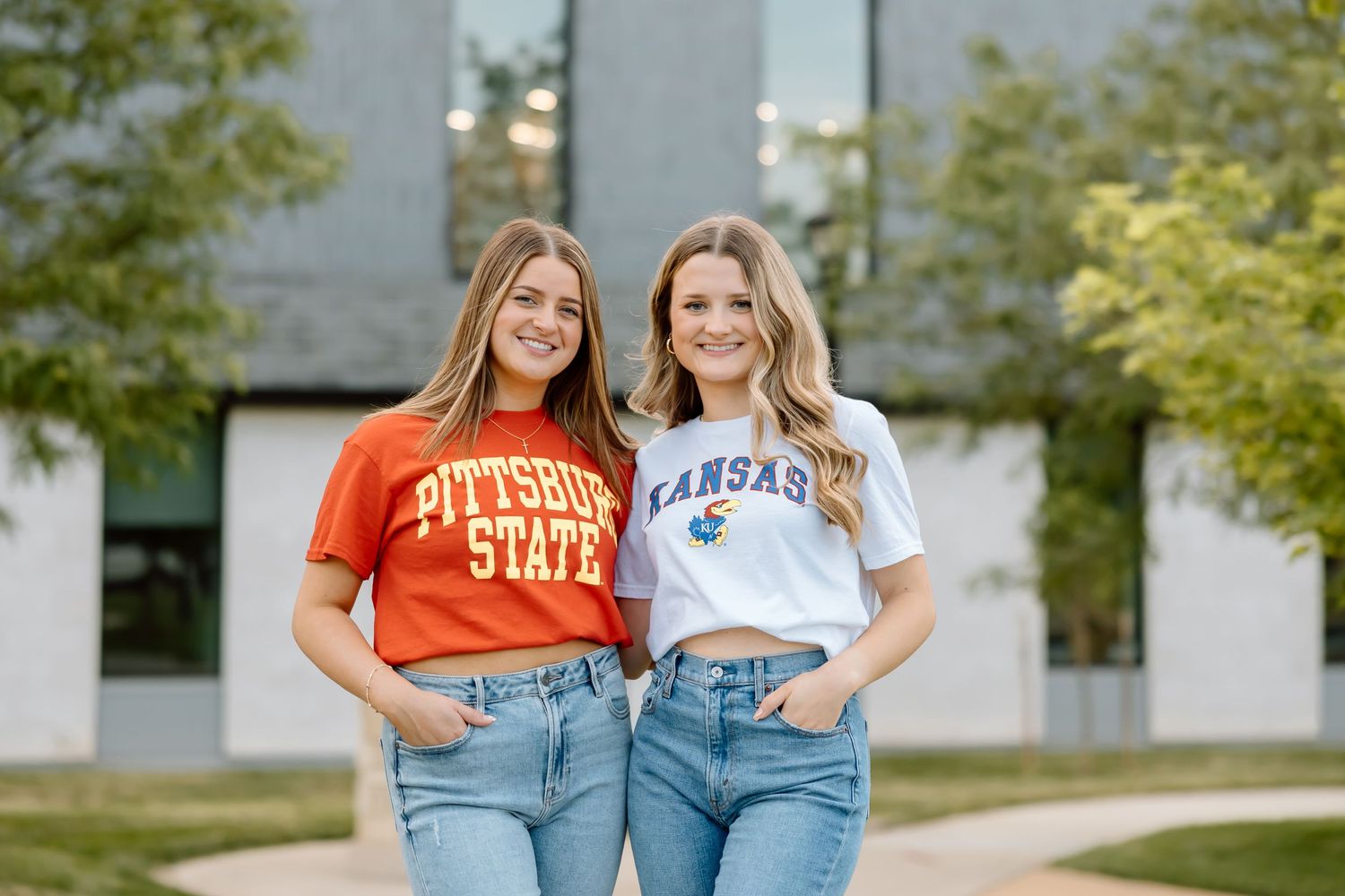Two people wearing orange college t-shirts and blue jeans pose together outdoors near trees and buildings.