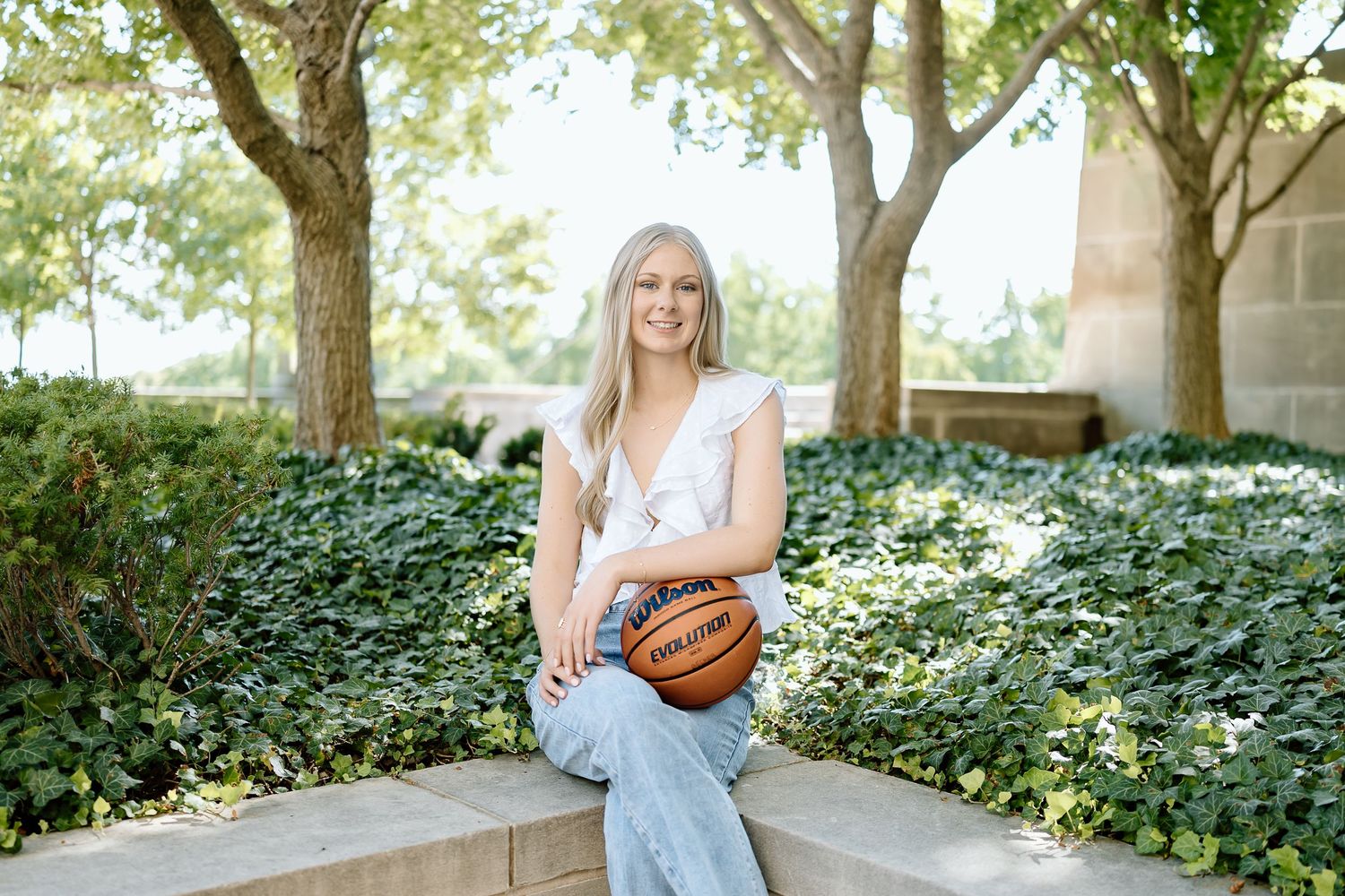 Basketball player poses outdoors in casual denim outfit against green foliage background.