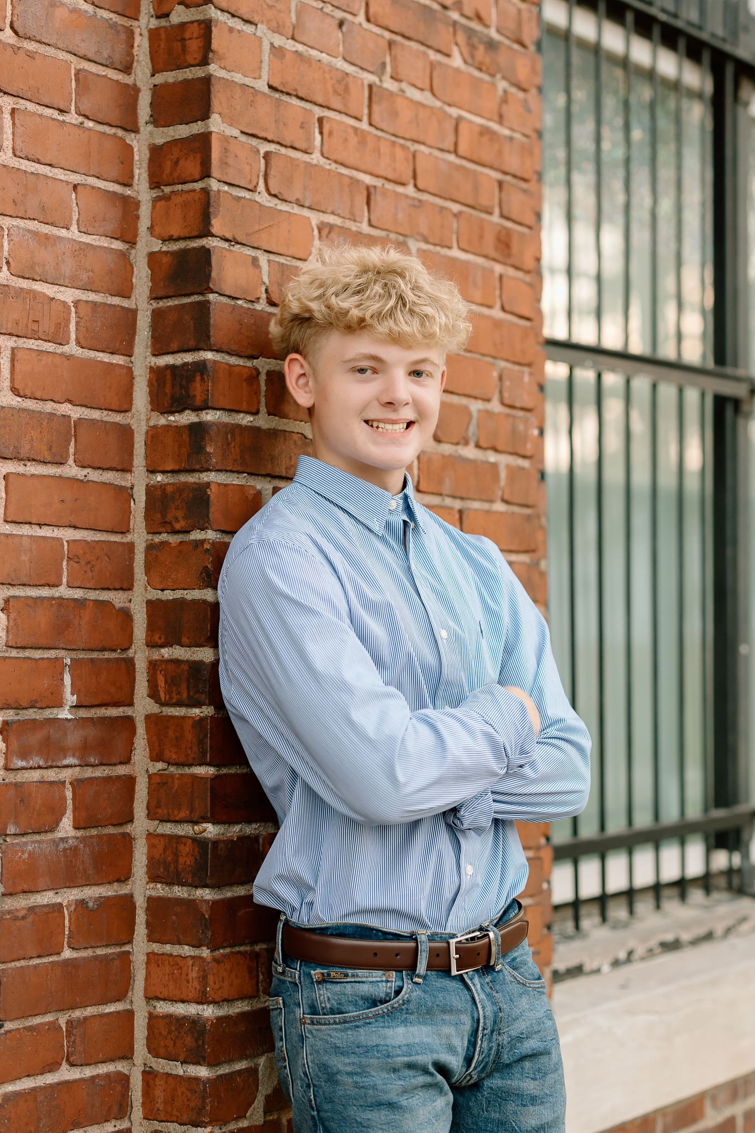 A person in a light blue button-down shirt and jeans leans against a red brick wall near a window.
