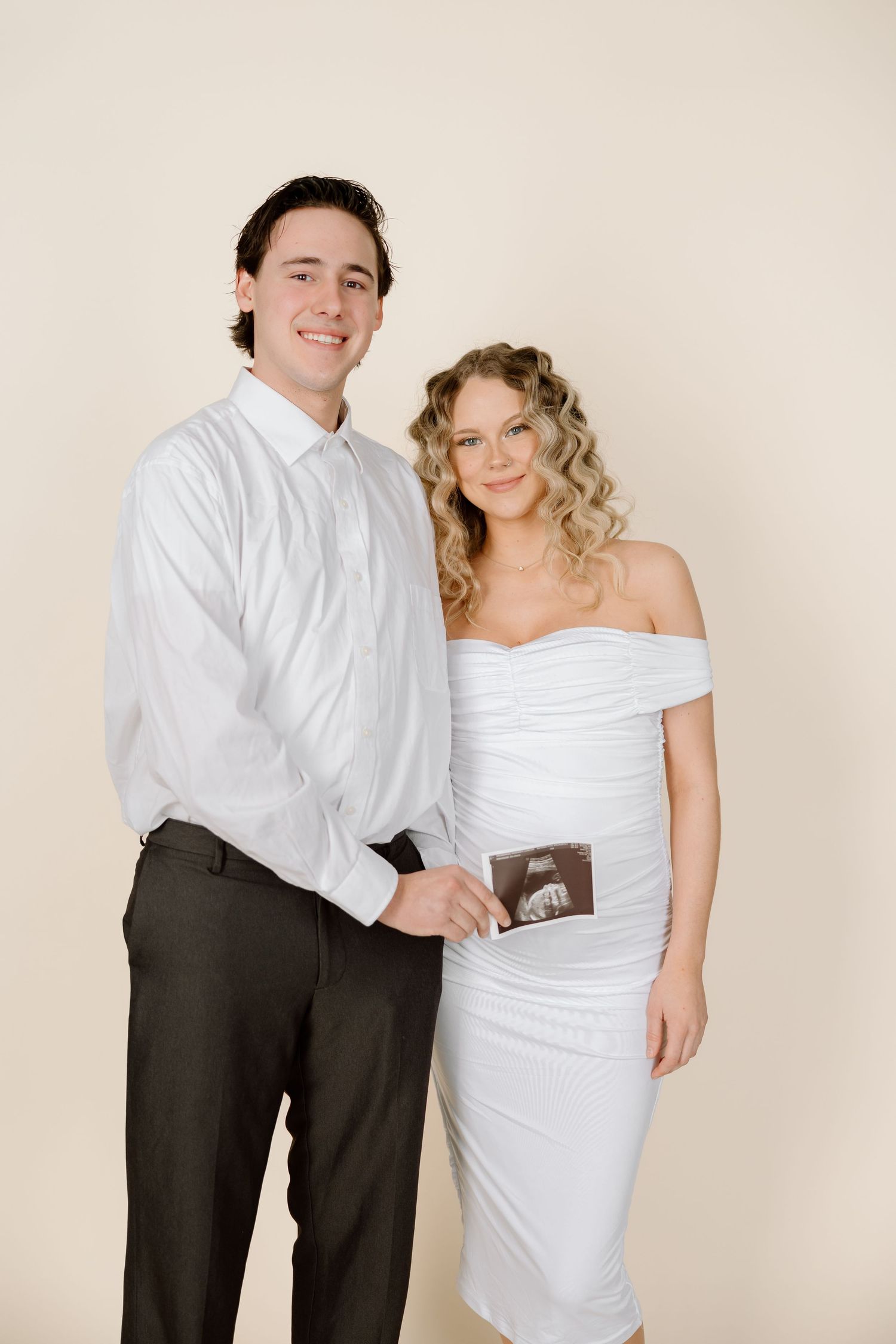 A couple in formal attire poses for a pregnancy announcement photo series against a neutral background.