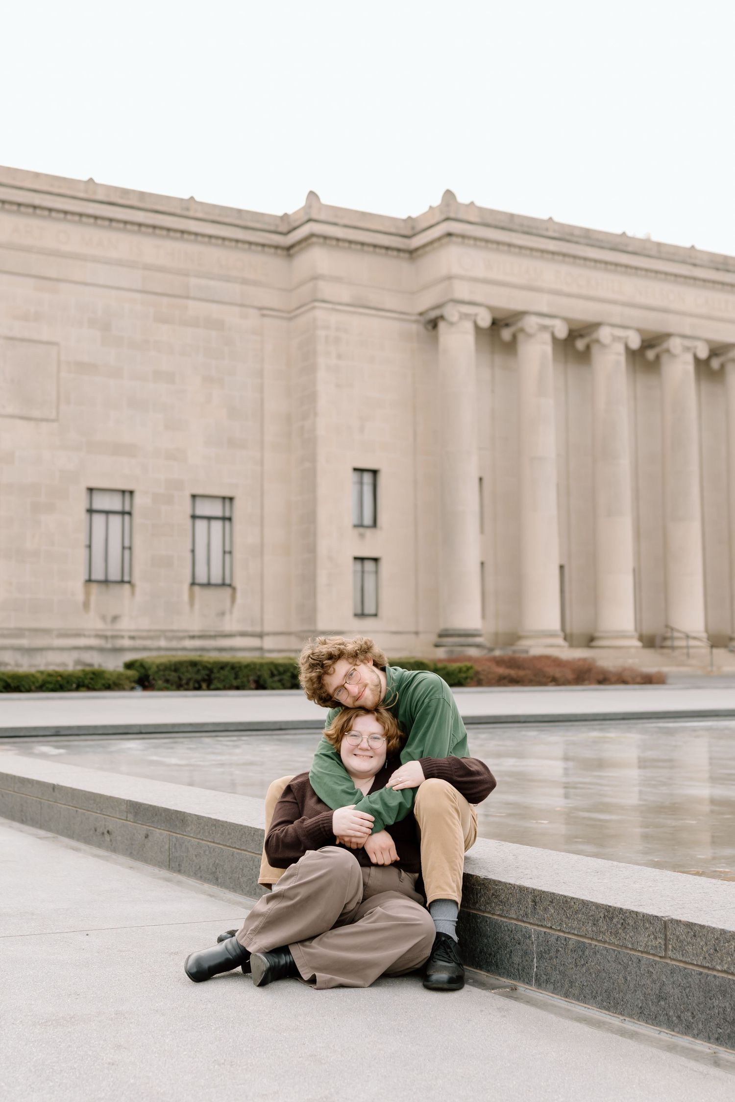A couple embraces and plays playfully on a stone ledge outside a grand stone building.