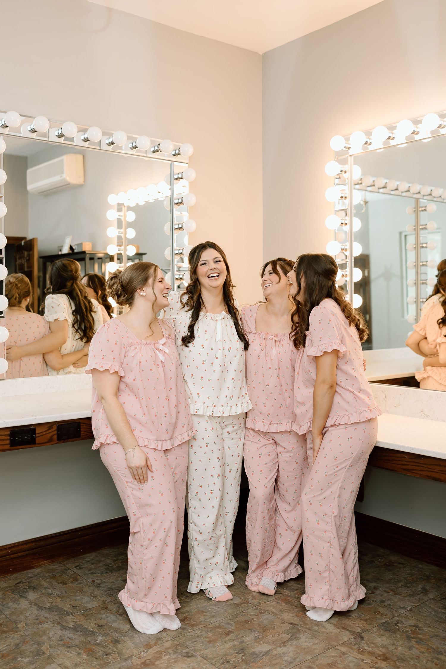 Group wearing matching pink and white pajama sets gather in front of lighted vanity mirrors in a dressing room.